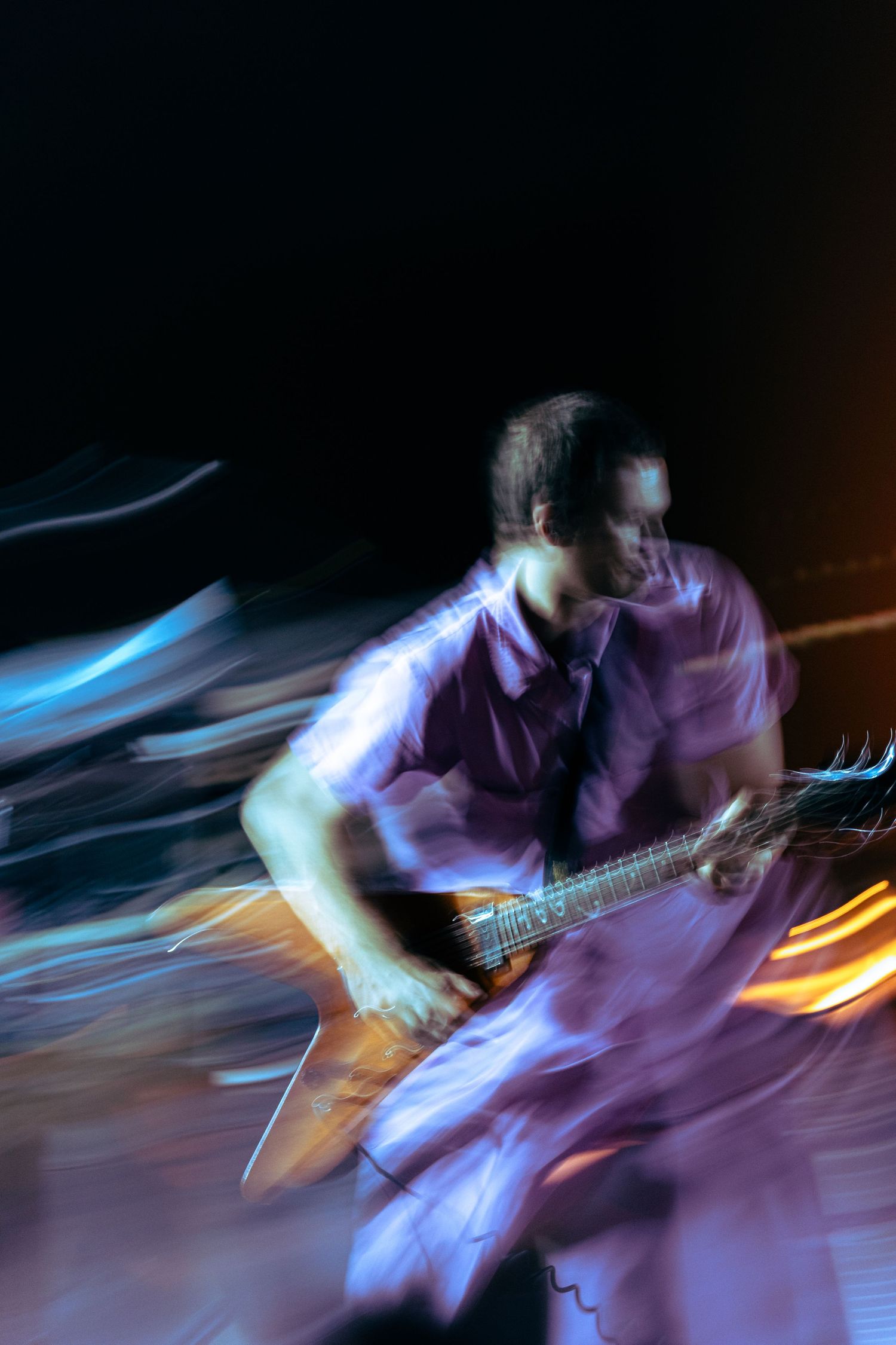 Motion blur concert photo of a guitarist in a purple shirt playing electric guitar under colorful stage lights.