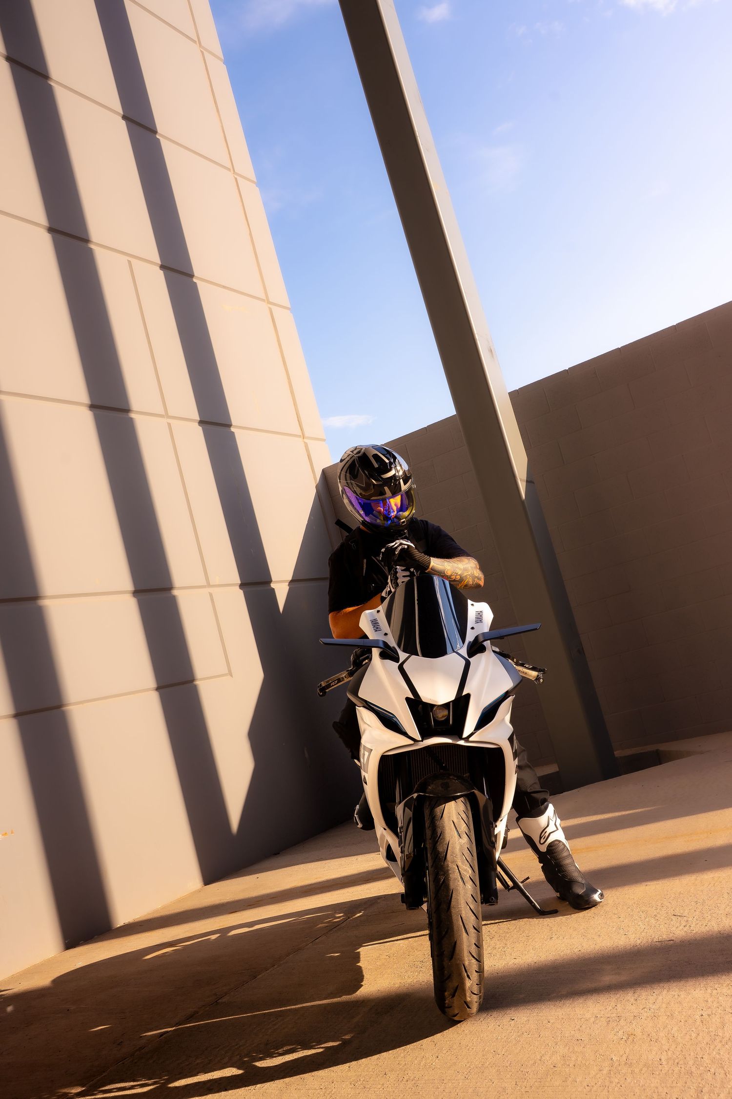 Biker in purple-tinted helmet seated on white sport motorcycle against concrete wall in sunlight.