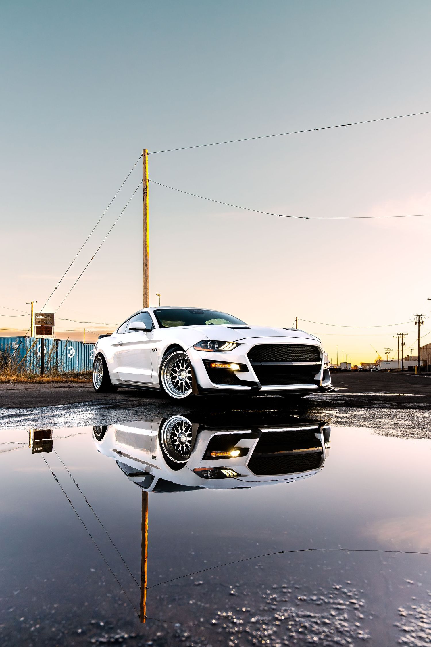 White Ford Mustang reflects perfectly in a puddle at golden hour near utility poles at sunset.