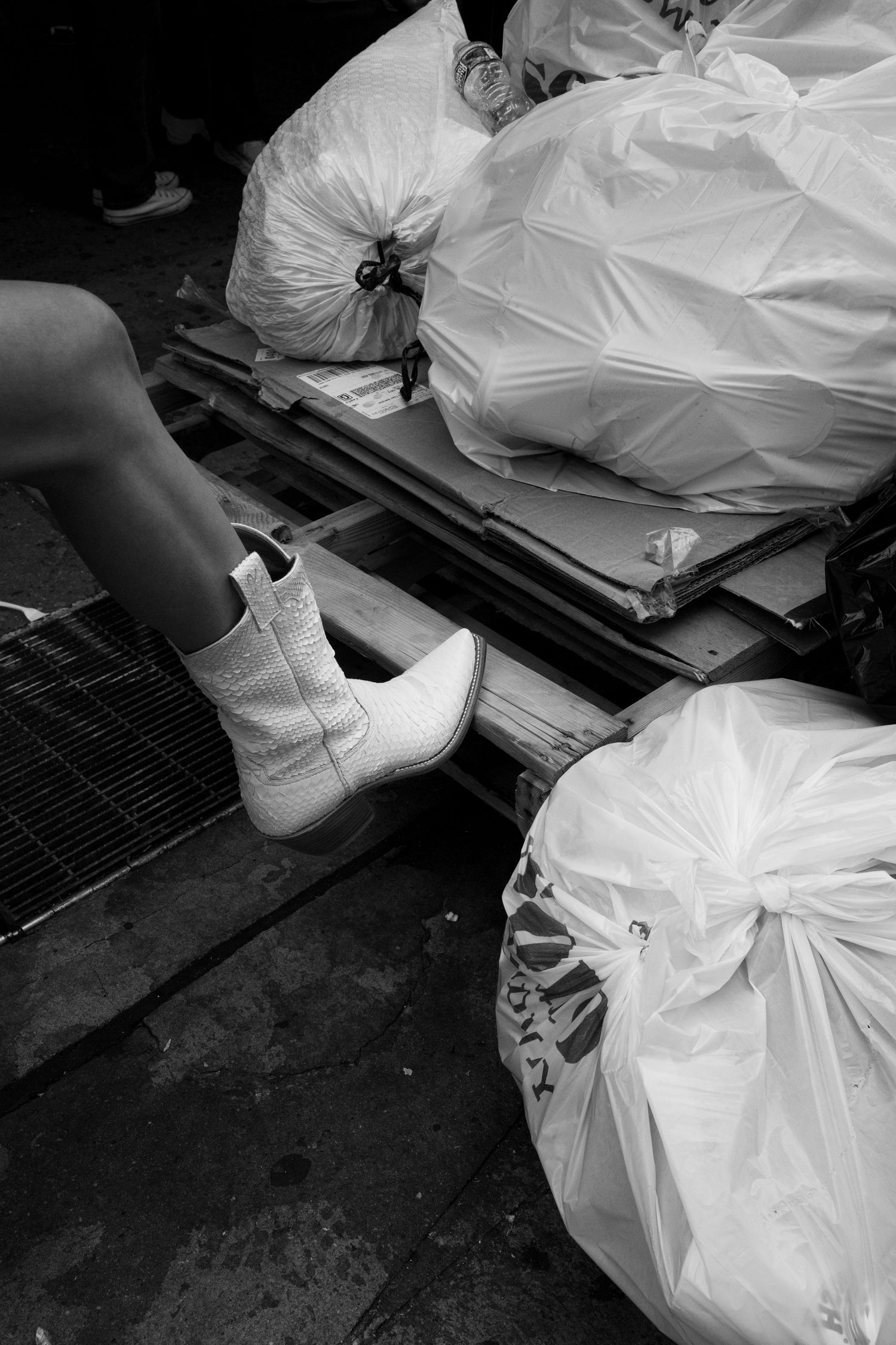 Black and white photo of white cowboy boots beside trash bags on a city sidewalk.