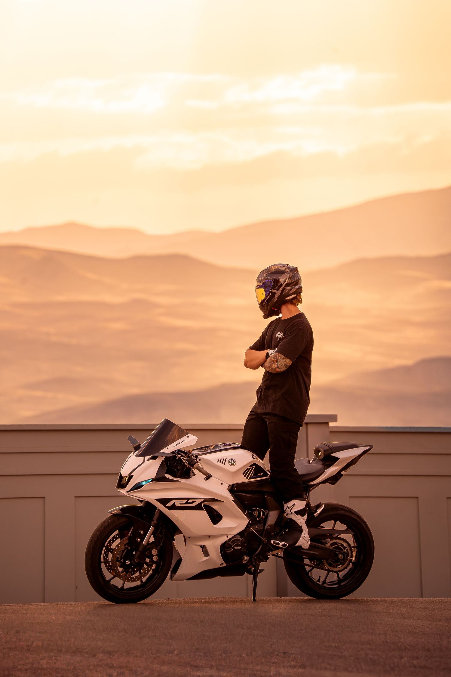 Helmeted rider with crossed arms standing on a white Yamaha R6 on a rooftop against a golden mountain sunset.
