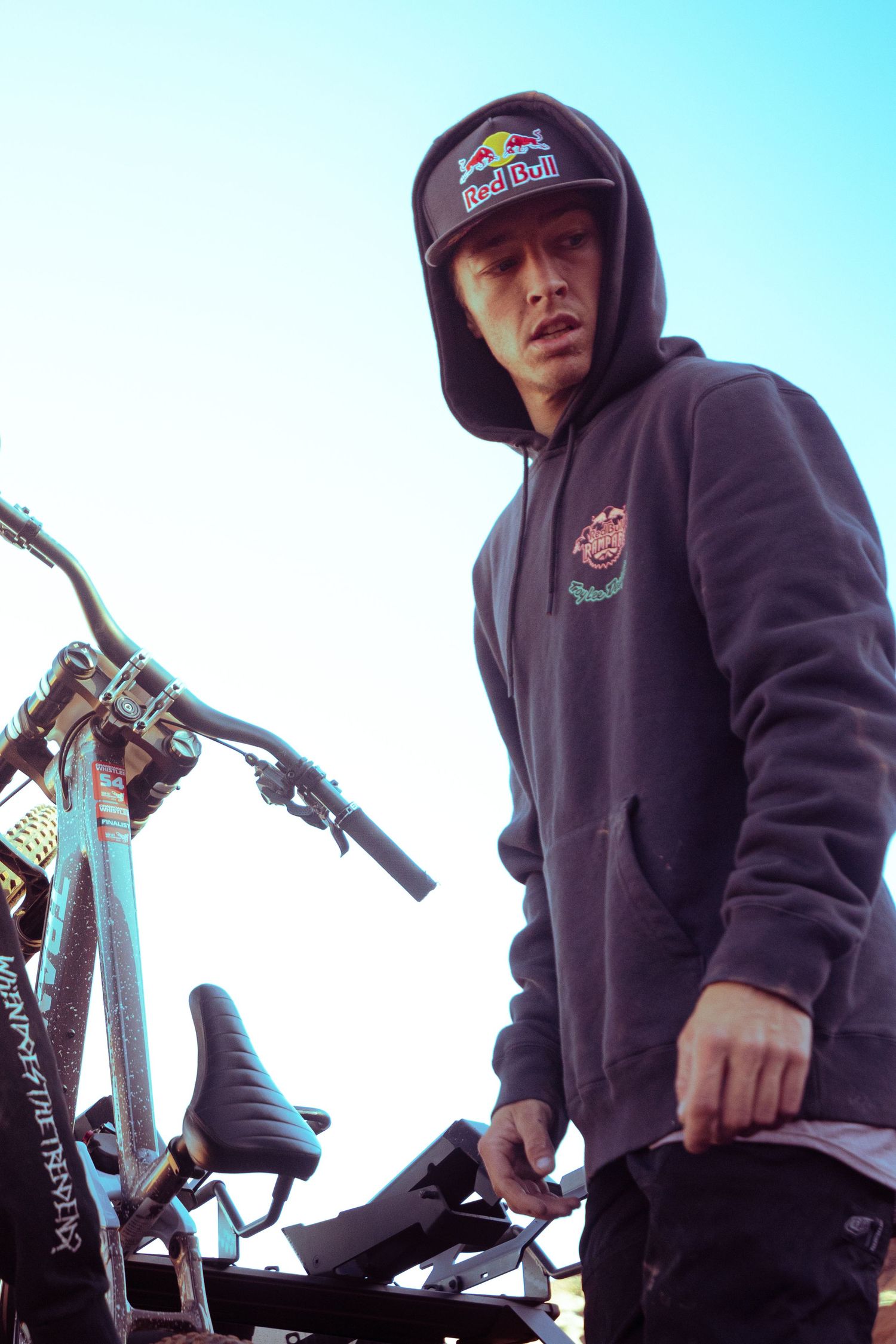 Young man in a Red Bull hoodie and cap standing beside a mountain bike against a bright sky.