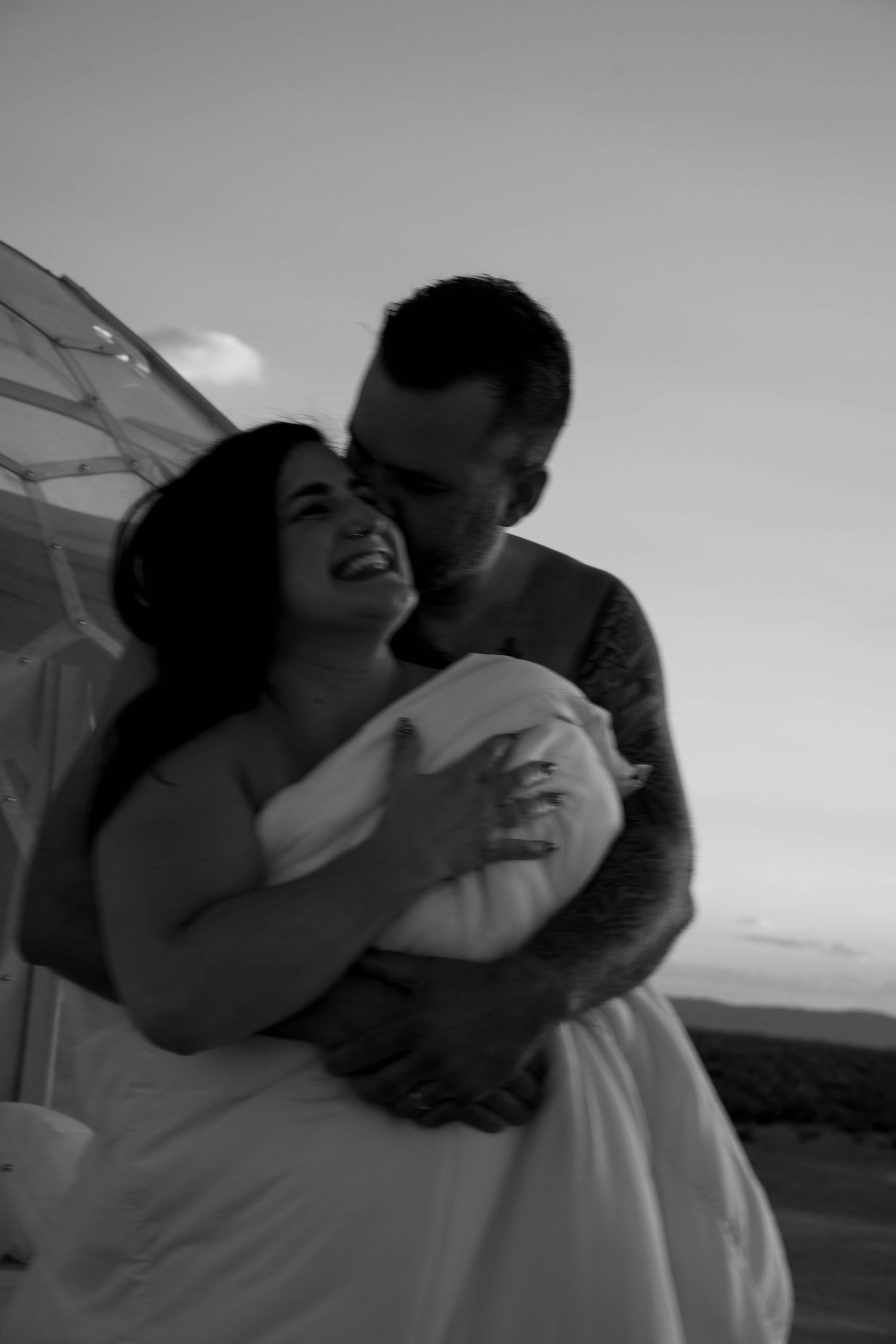 Black and white romantic photo of laughing bride being embraced by groom at sunset.