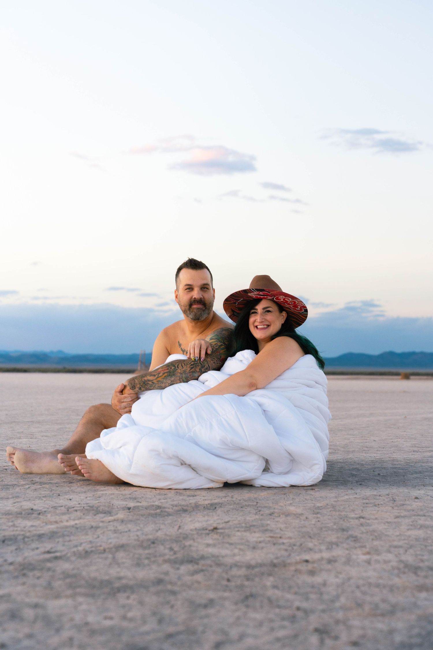 Couple wrapped in white blanket sitting on dry lake bed at dusk.