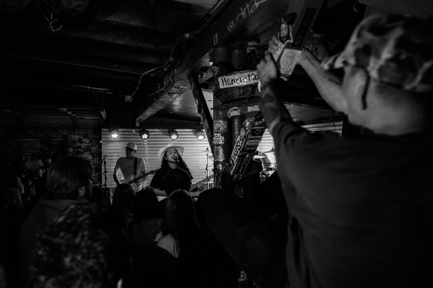 Black and white photo of a band performing live in a rustic indoor venue with audience members watching.