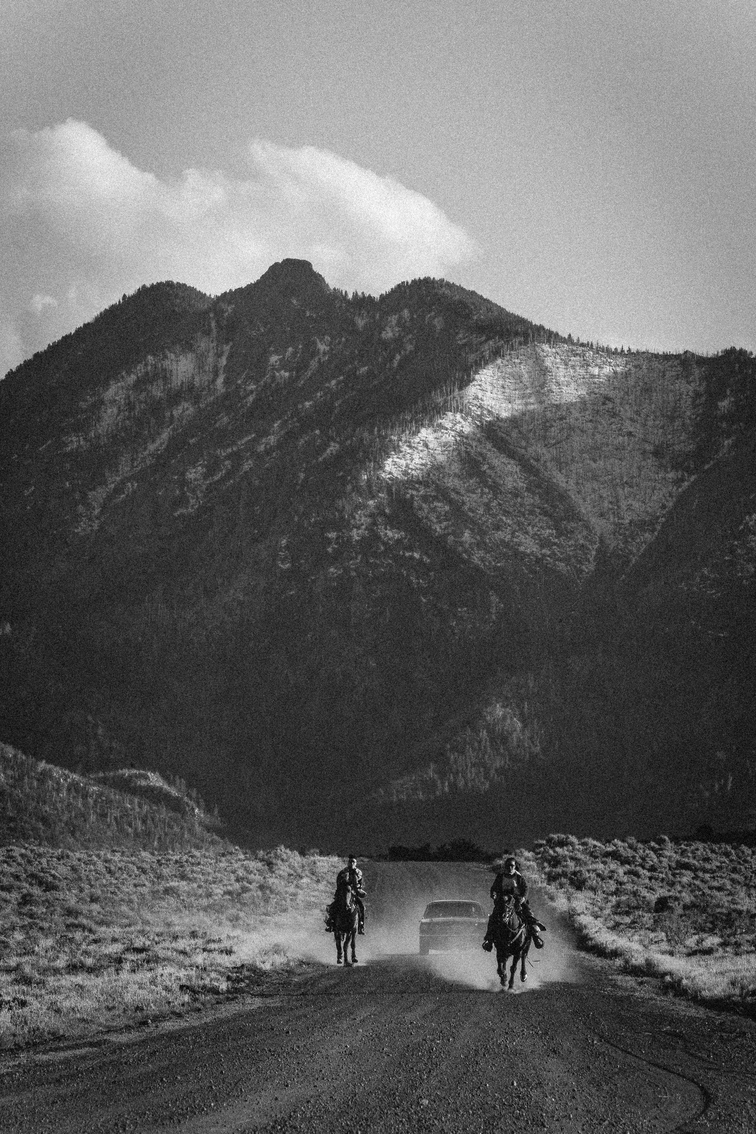 Black and white photo of two riders on horseback racing a car down a dusty mountain road.