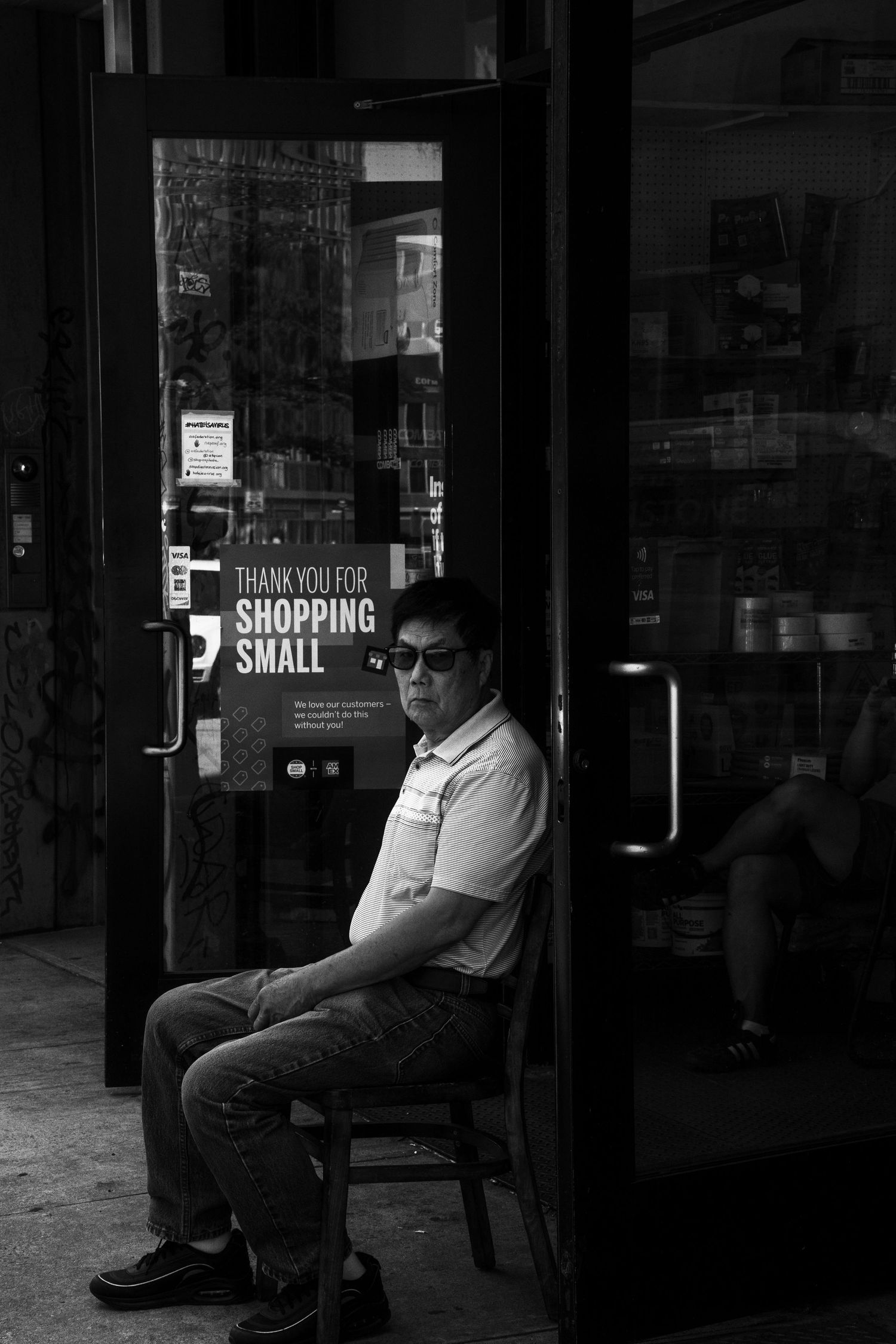 Black and white street photo of a man in sunglasses sitting outside a shop with Thank You For Shopping Small sign.