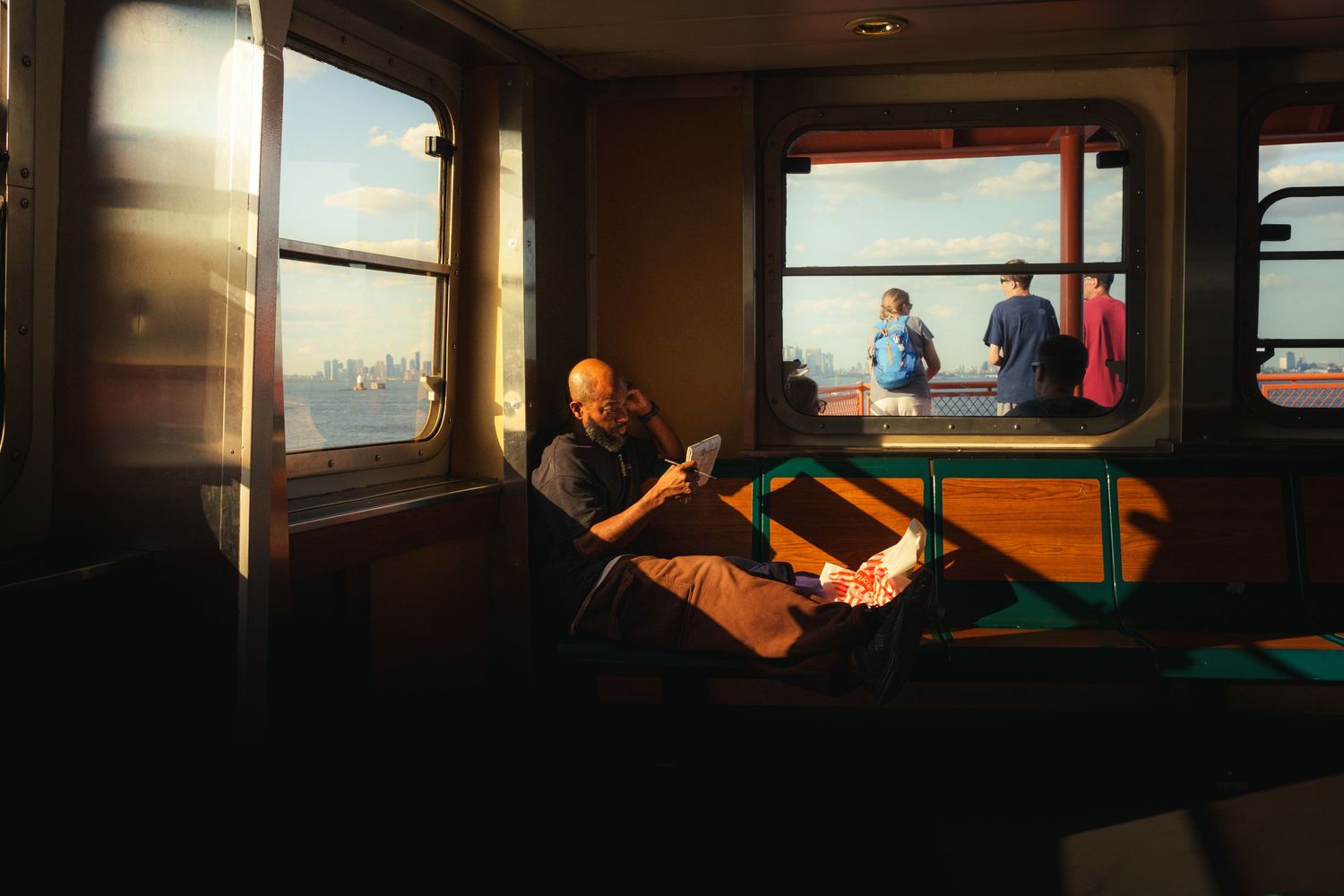 Man relaxing in warm golden sunlight inside a ferry boat cabin with city views through the windows.