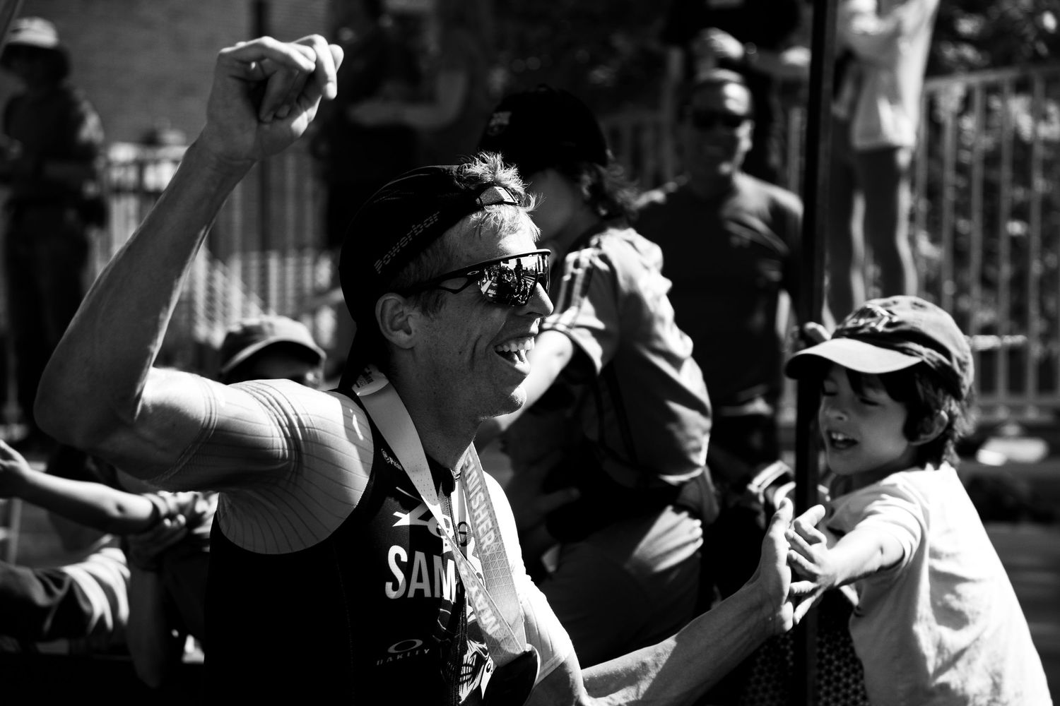 Black and white photo of a triathlete celebrating with a young fan at an Ironman race finish area.