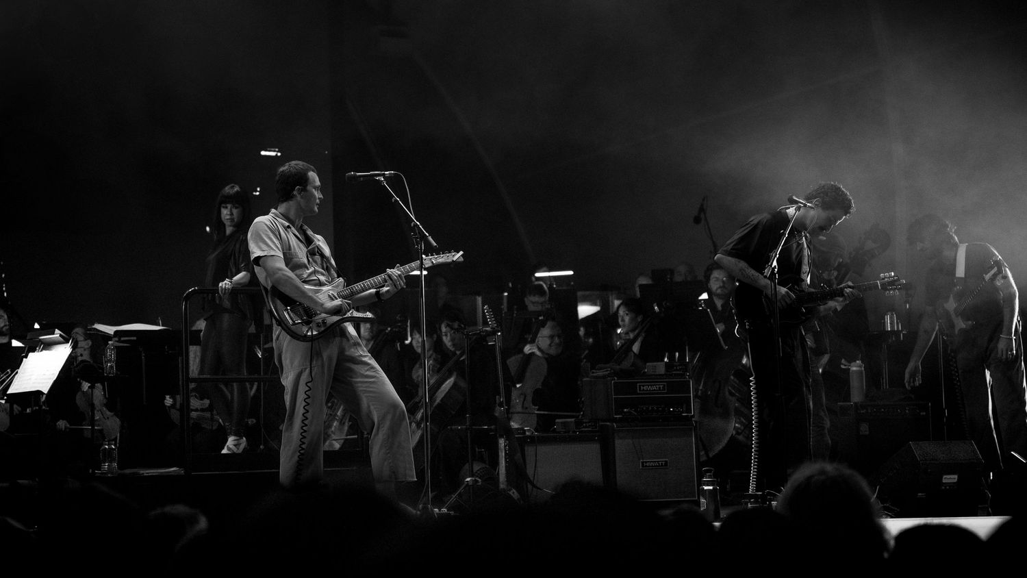 Black and white photo of two musicians performing on stage, one playing guitar and one playing saxophone.