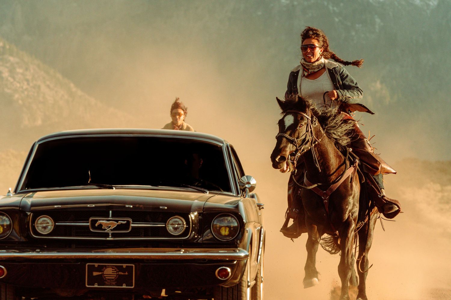 A woman on horseback gallops alongside a classic Ford Mustang in a dramatic dusty mountain landscape.