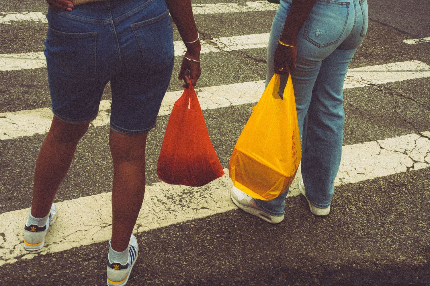 Two women carrying colorful plastic bags crossing a zebra crosswalk on a city street.