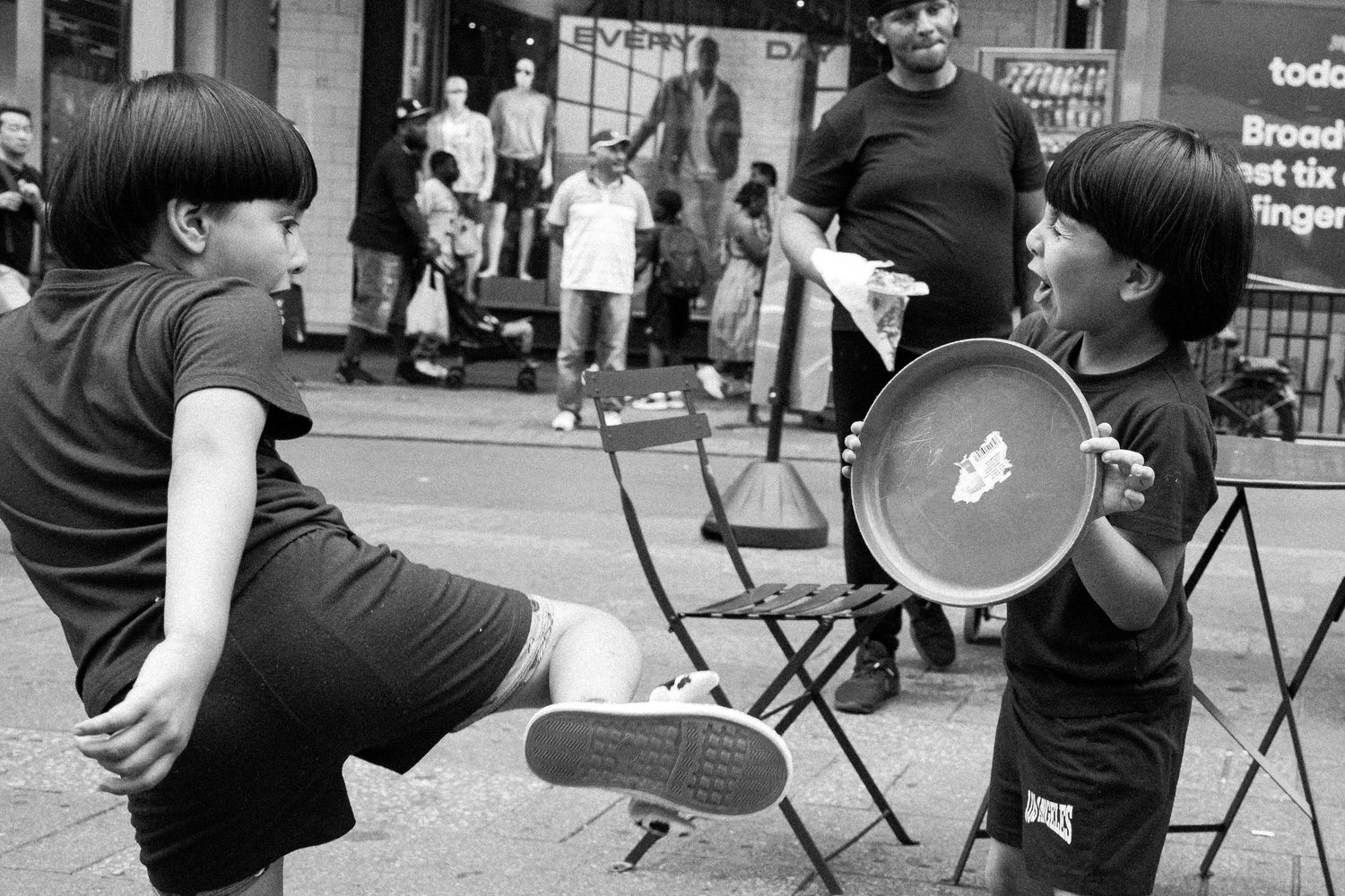 Black and white photo of two children playing with a tambourine on a busy city street.
