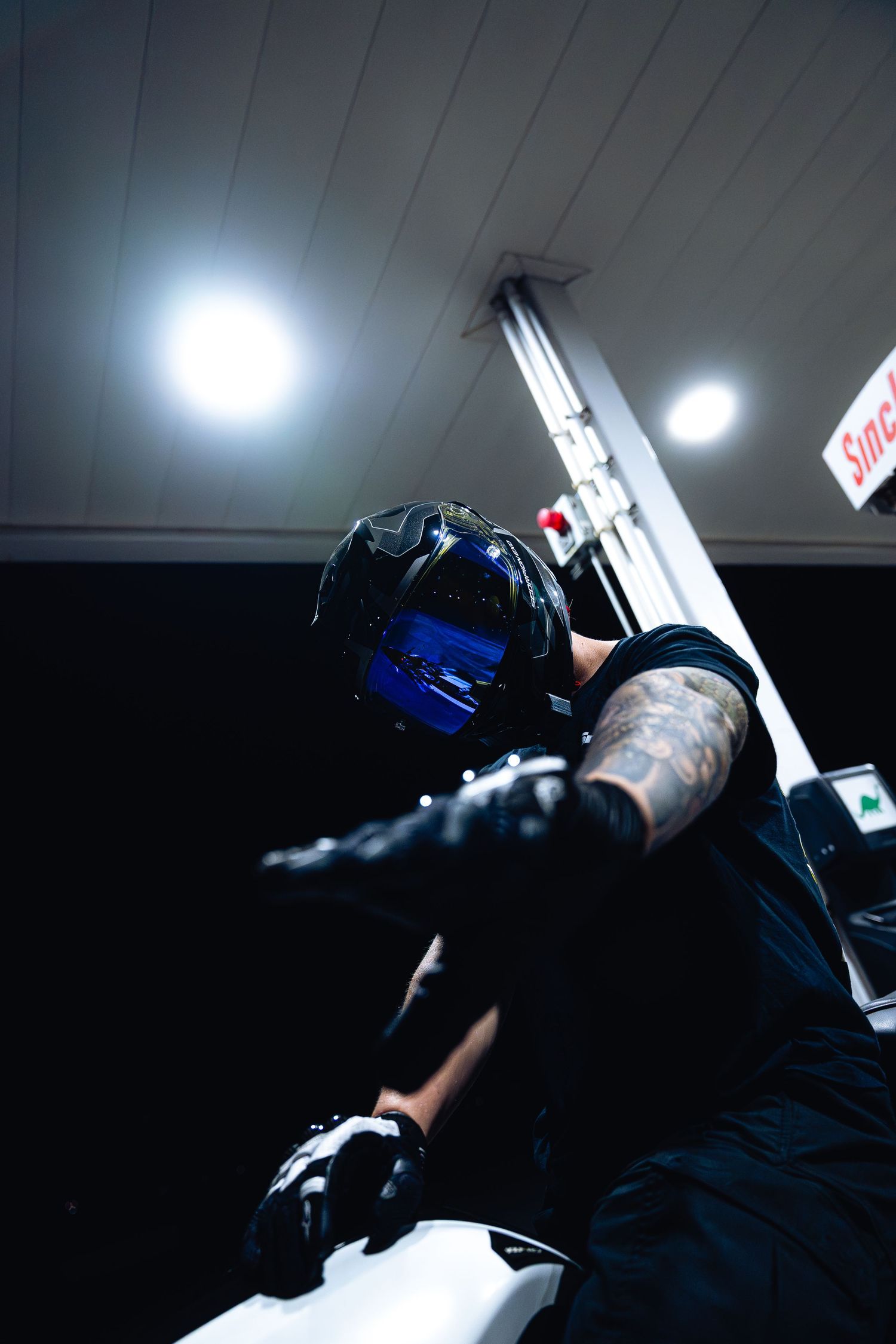 Low angle view of tattooed biker in black helmet with blue visor leaning forward on motorcycle at gas station.