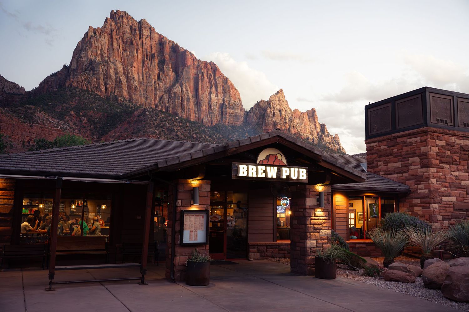 Zion Brew Pub exterior at sunset with dramatic red rock canyon cliffs of Zion National Park behind it.