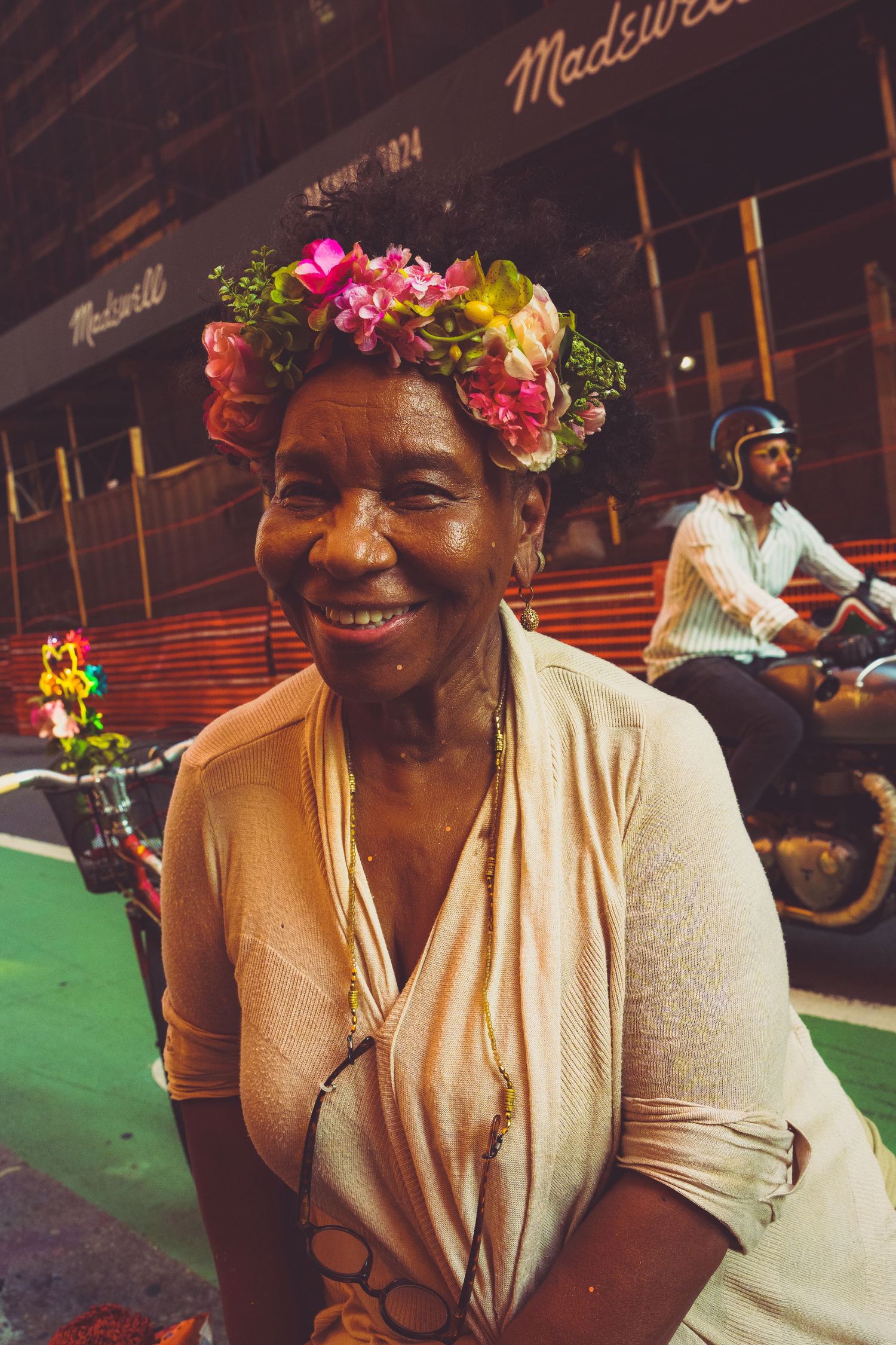 Smiling elderly woman wearing a colorful floral crown at a city street event near a Madewell store.