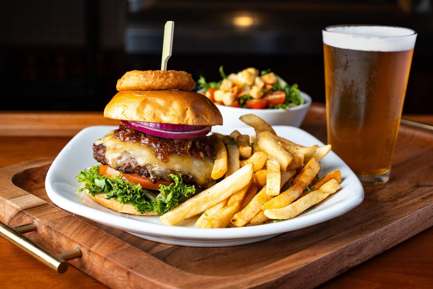 Juicy cheeseburger with fries and a cold beer served on a wooden board at a casual dining restaurant.