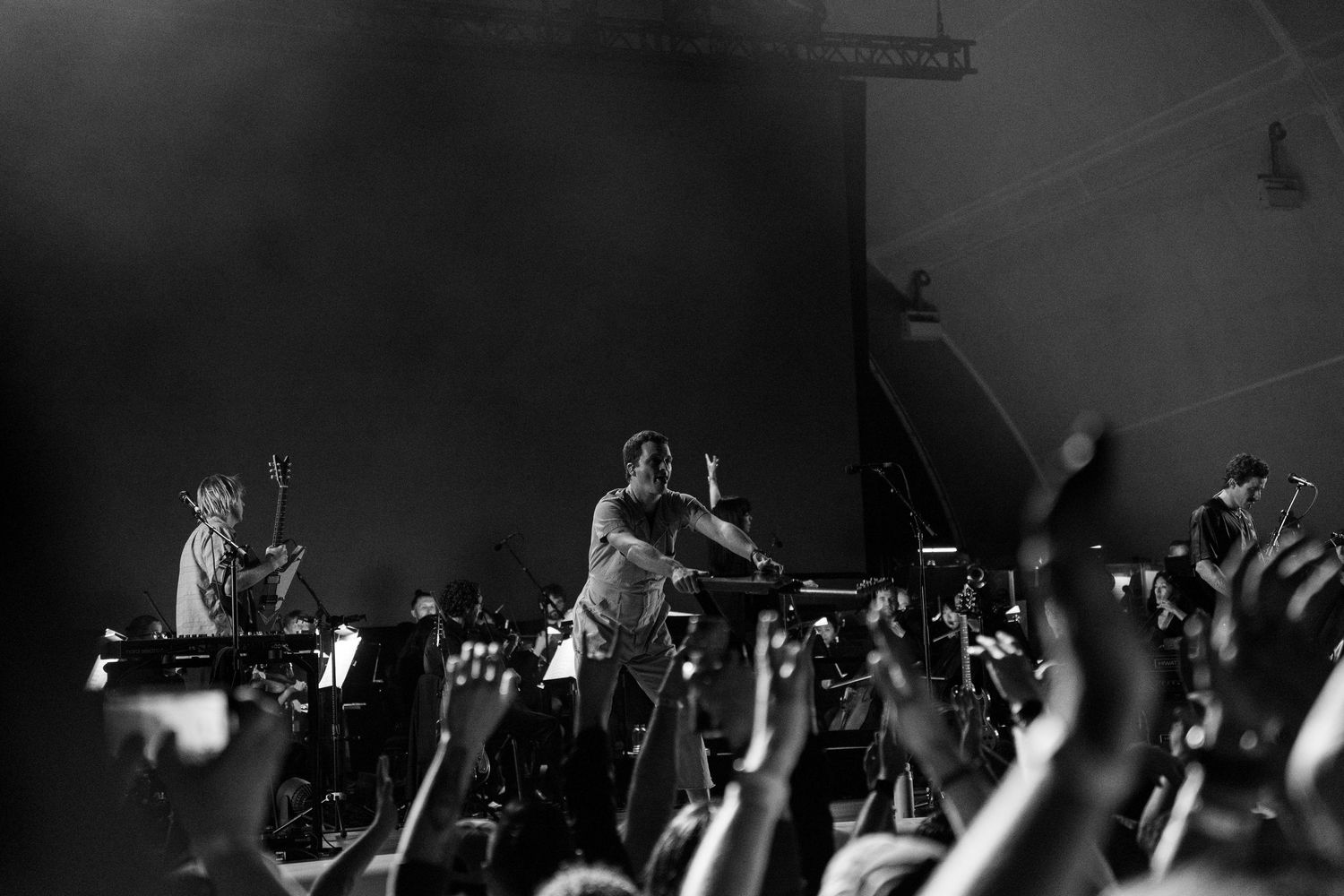 Black and white wide shot of concert performers on stage with crowd raising hands in the foreground.