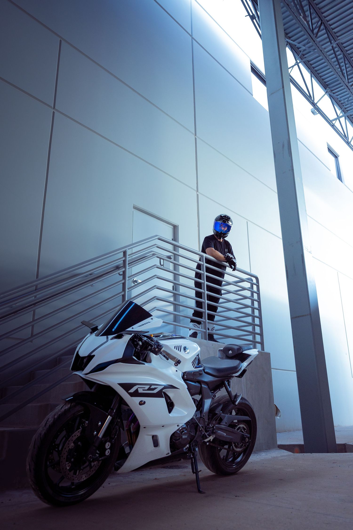 Helmeted rider standing on stairs beside white Yamaha R7 sport motorcycle in blue-toned garage.