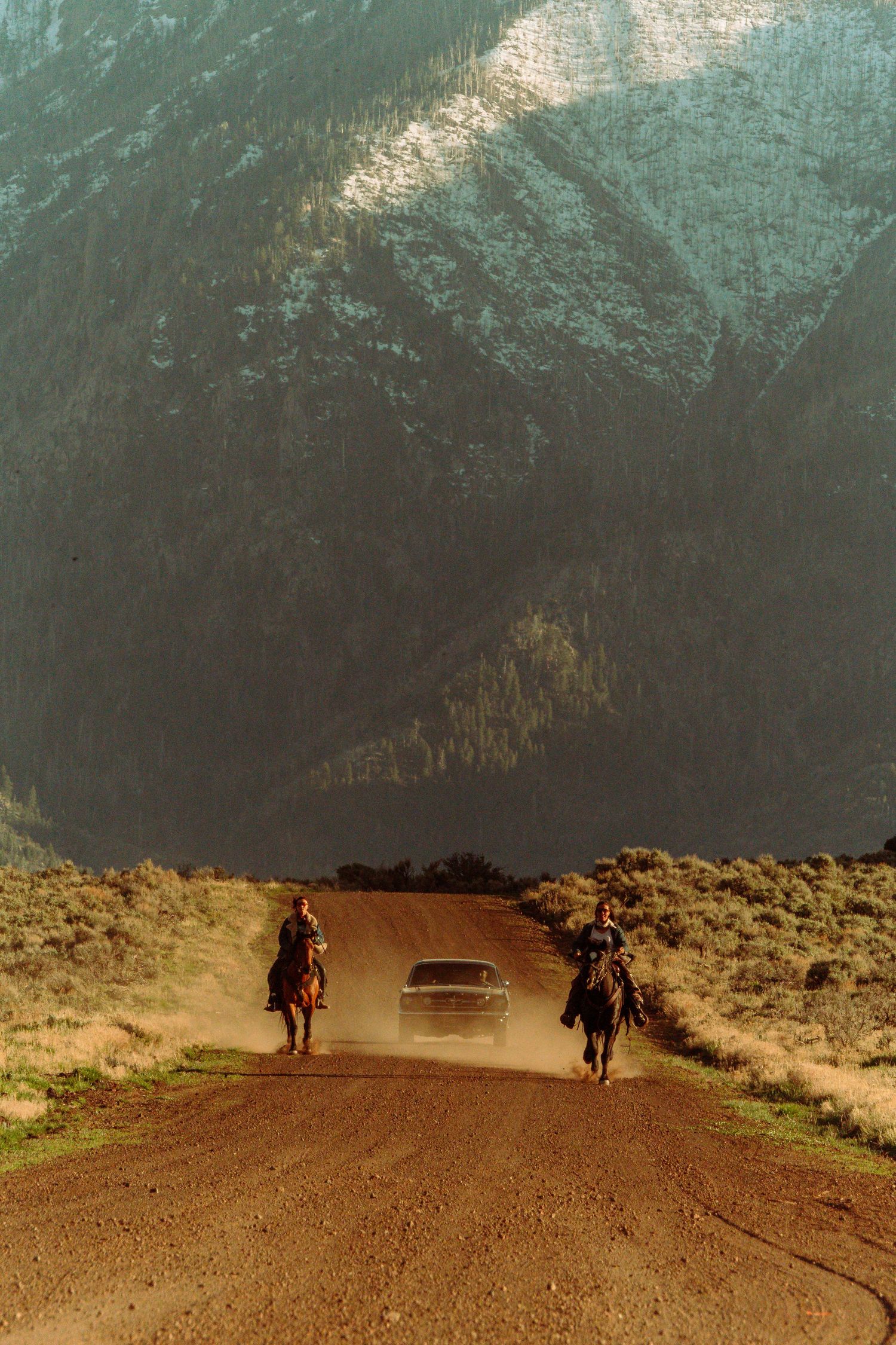 Two riders on horseback flank a dusty dirt road with a car approaching, set against dramatic mountain scenery.