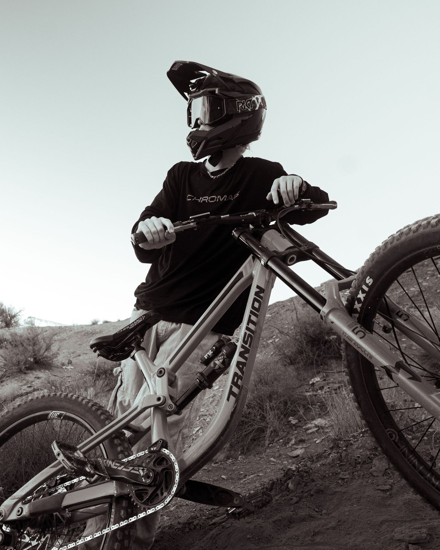 Black and white close-up of helmeted rider on Transition mountain bike on rugged dirt trail.