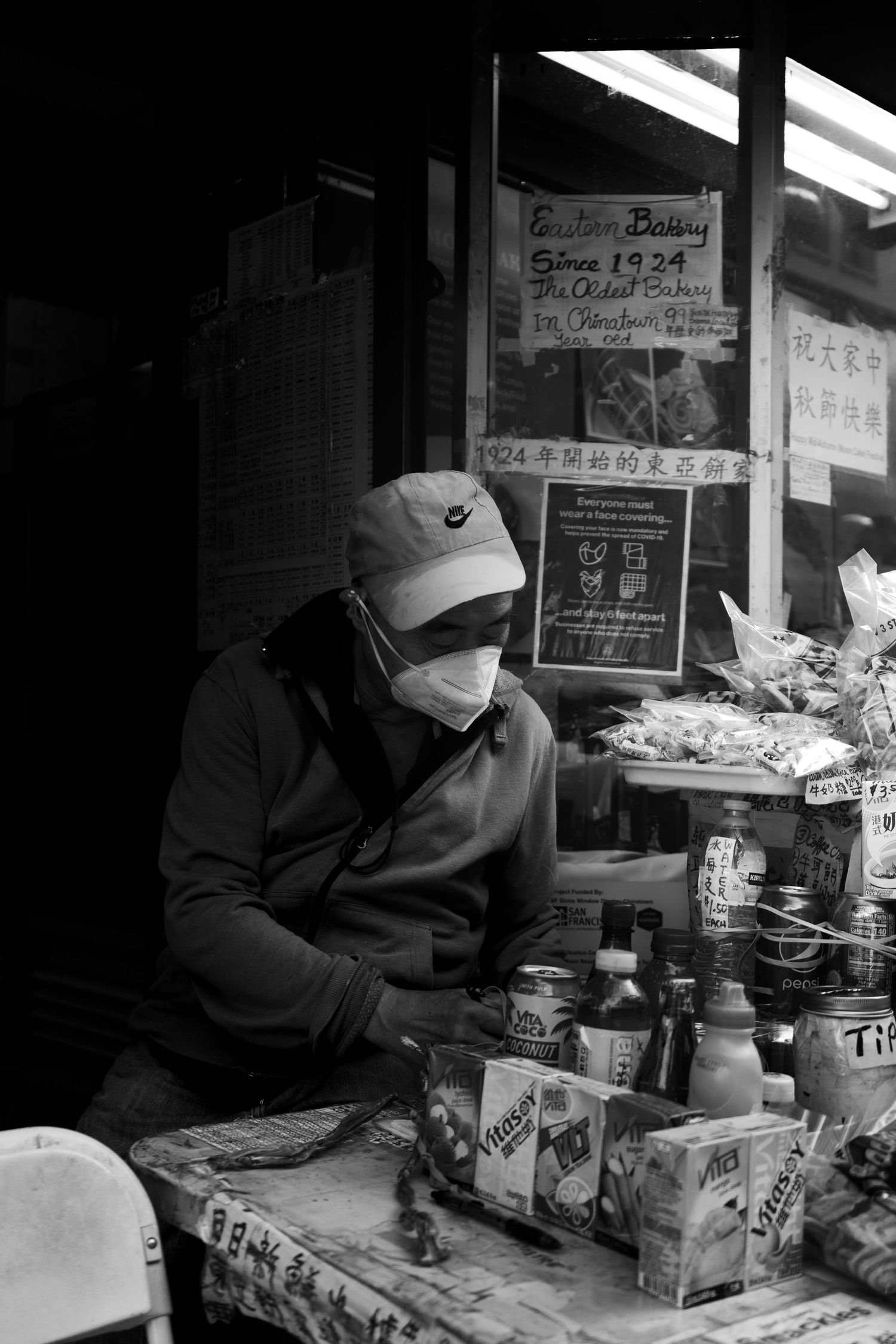 A masked vendor sits at a cluttered market stall with Chinese signage and various goods in black and white.