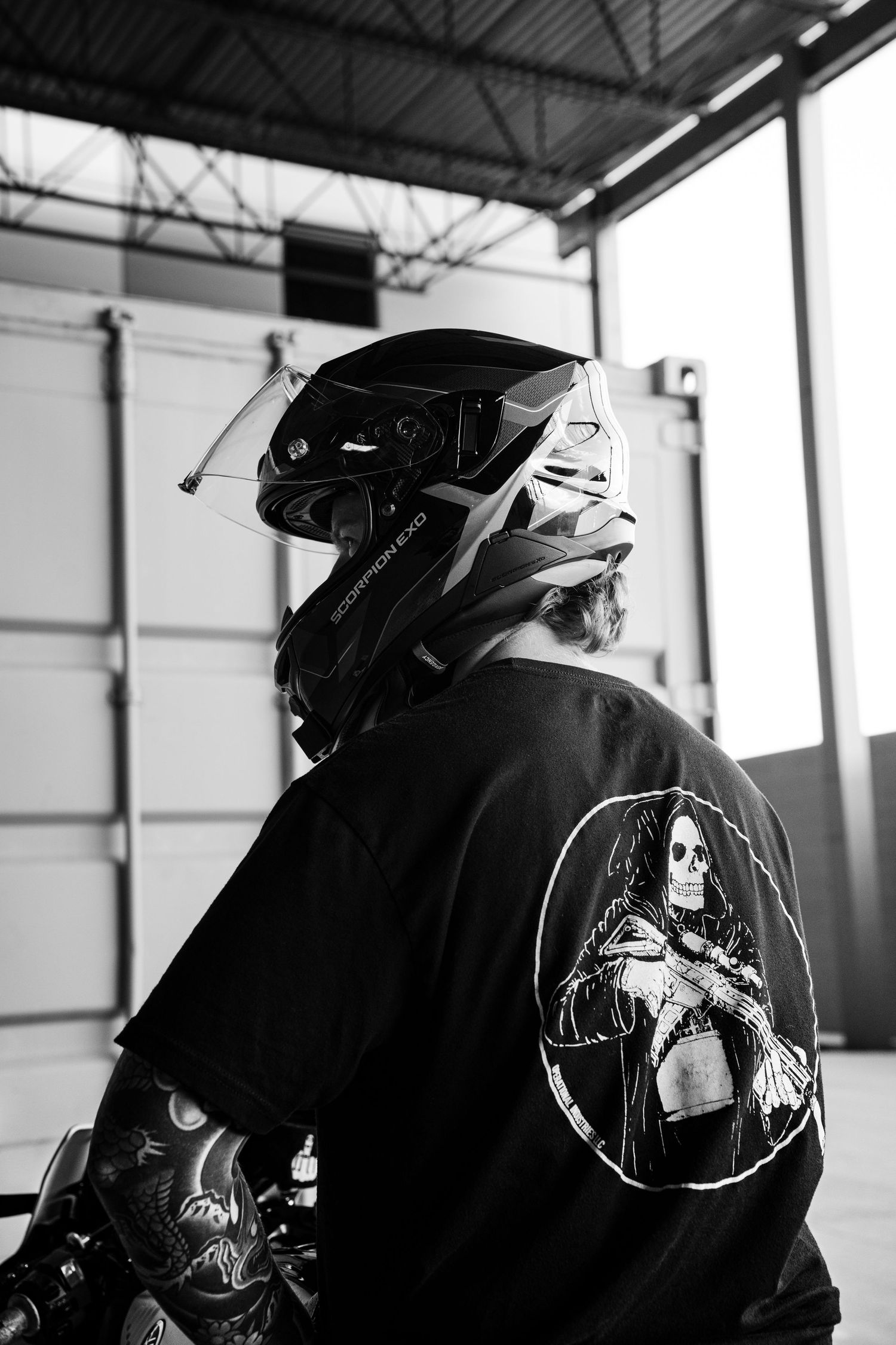 Black and white rear view of tattooed rider in skull graphic tee and helmet inside a garage.