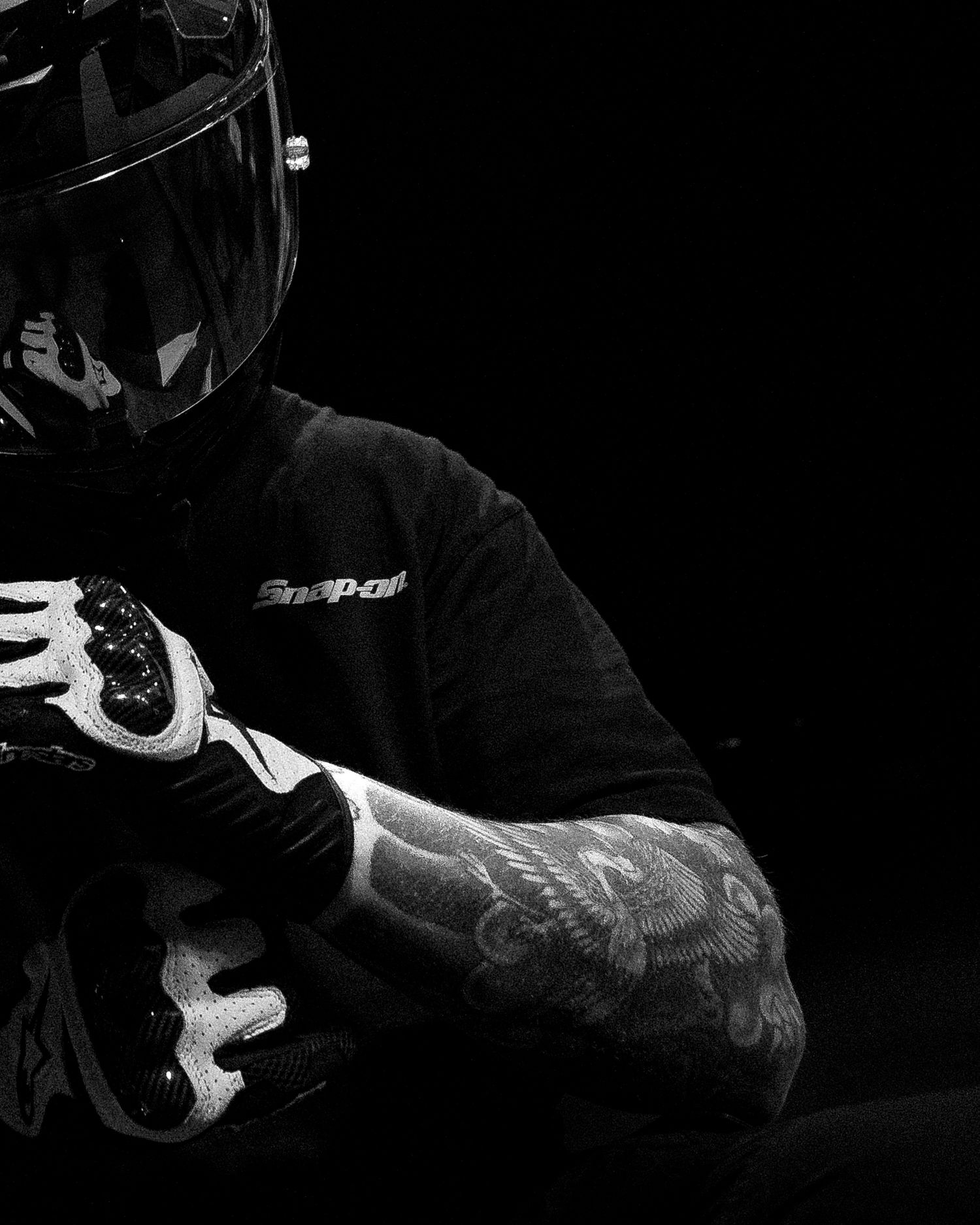 Black and white portrait of tattooed motorcyclist in Snap-on gear and full-face helmet.