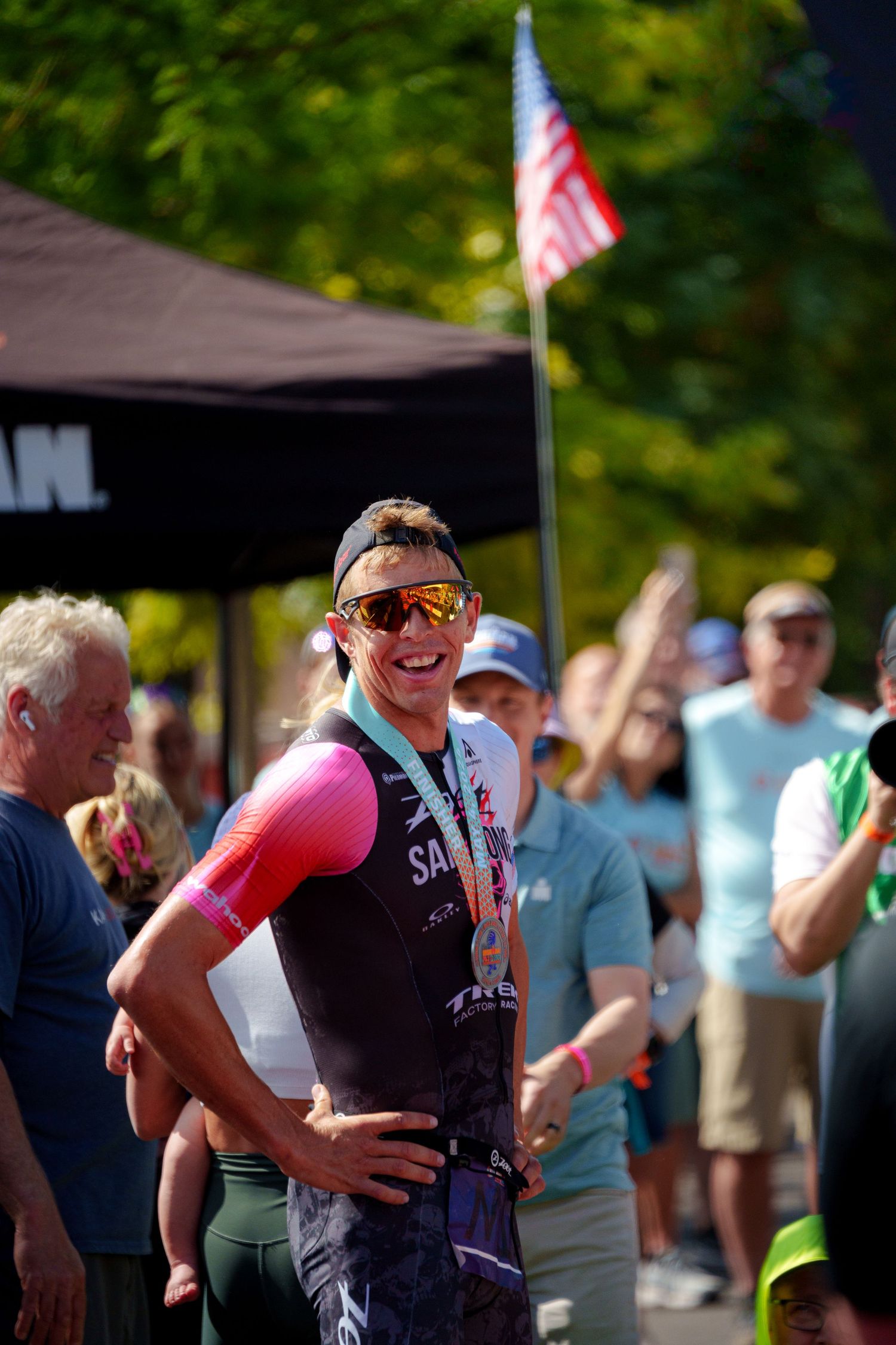 Smiling male triathlete wearing sunglasses and a finisher medal celebrates after completing an Ironman race.
