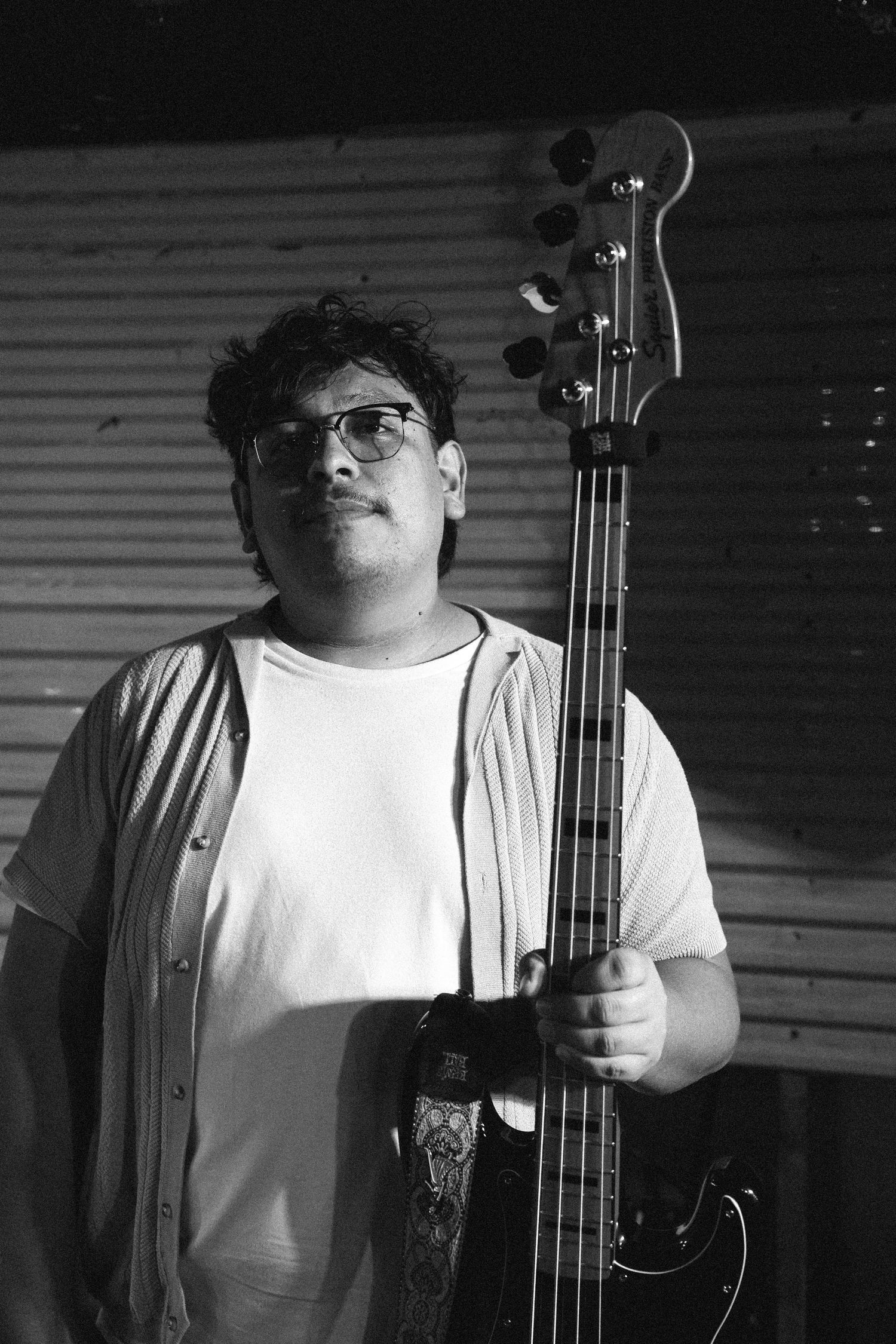 Black and white portrait of guitarist holding bass guitar upright, wearing glasses and casual shirt.