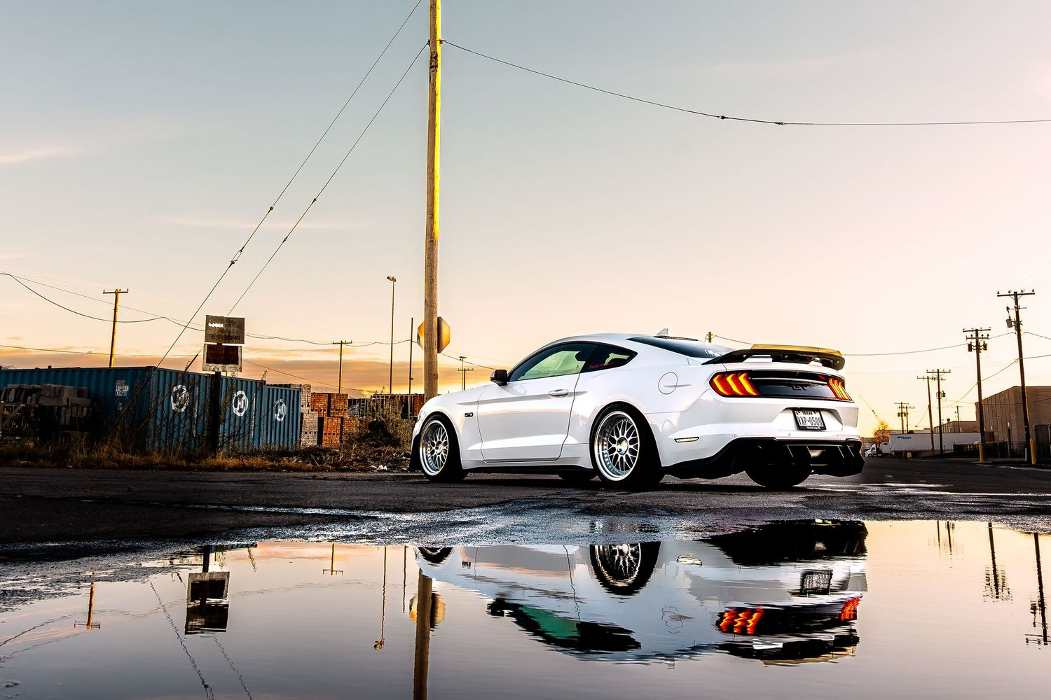 White Ford Mustang reflects in a puddle at golden hour near shipping containers and utility poles.
