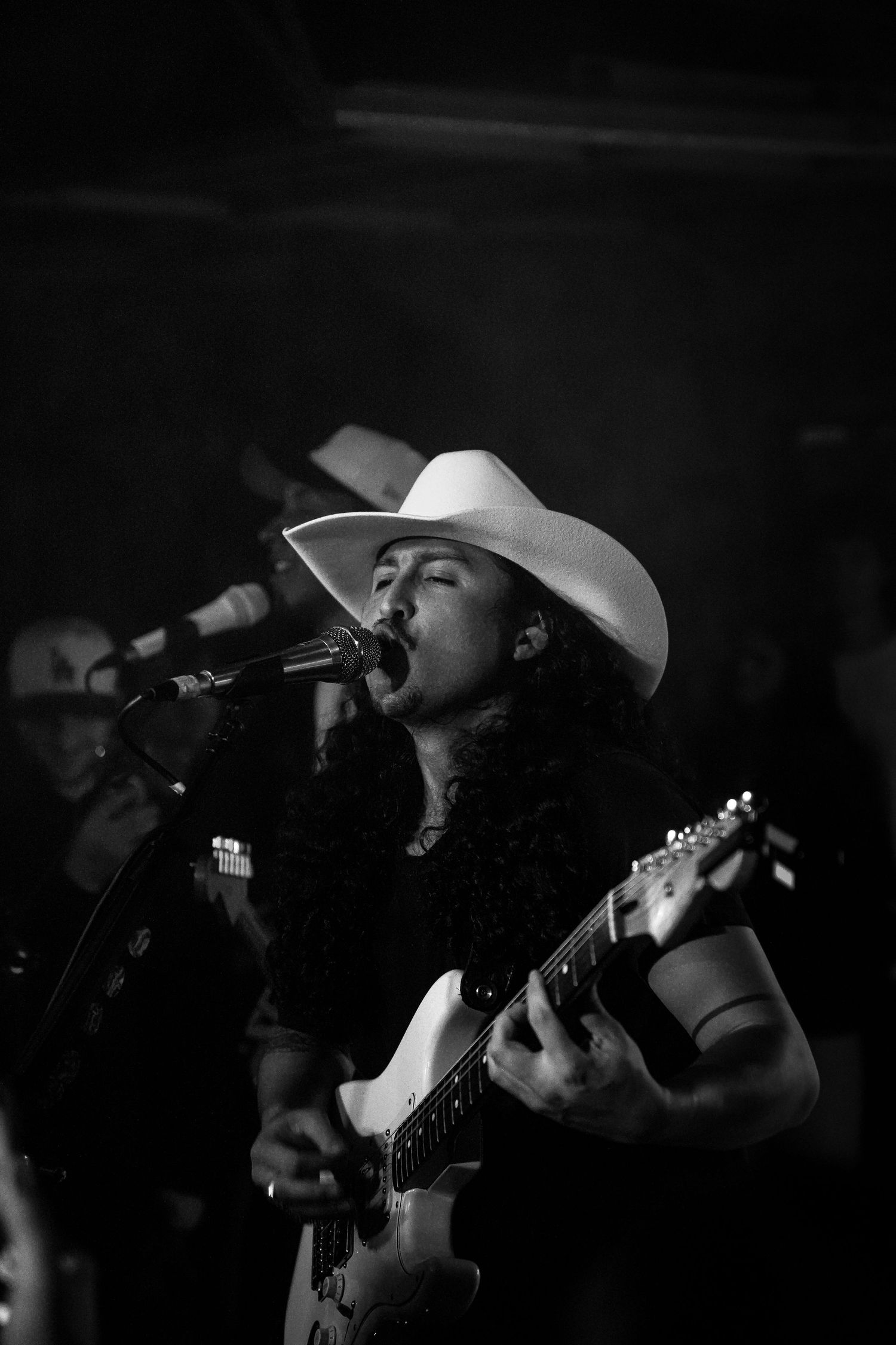 Black and white photo of a long-haired singer in a white cowboy hat playing guitar and singing into a mic.
