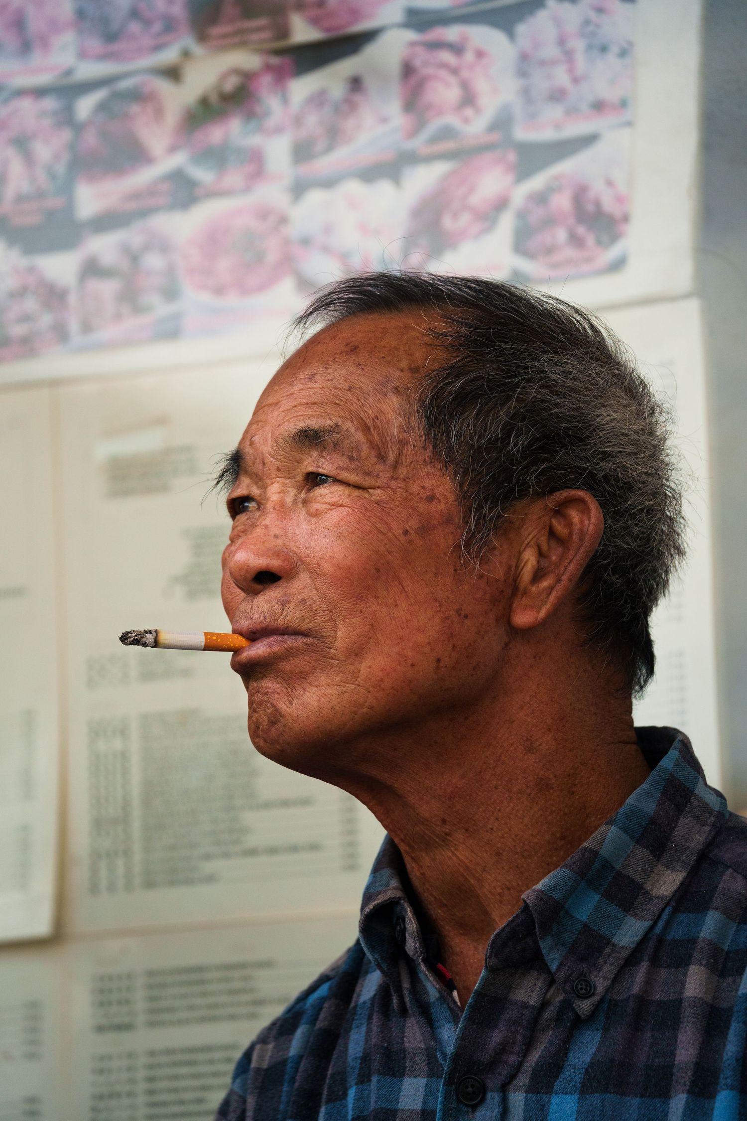 Elderly Asian man with a cigarette in his mouth standing in front of a poster on a wall.
