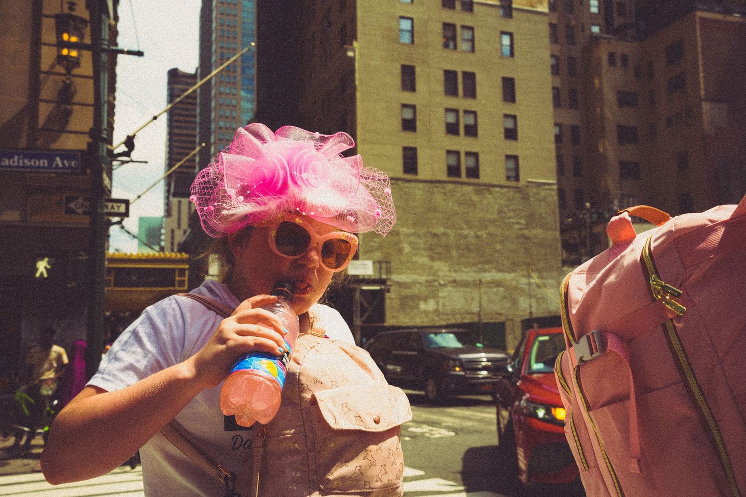 Woman in pink feather hat and sunglasses drinks beverage on busy New York City street.