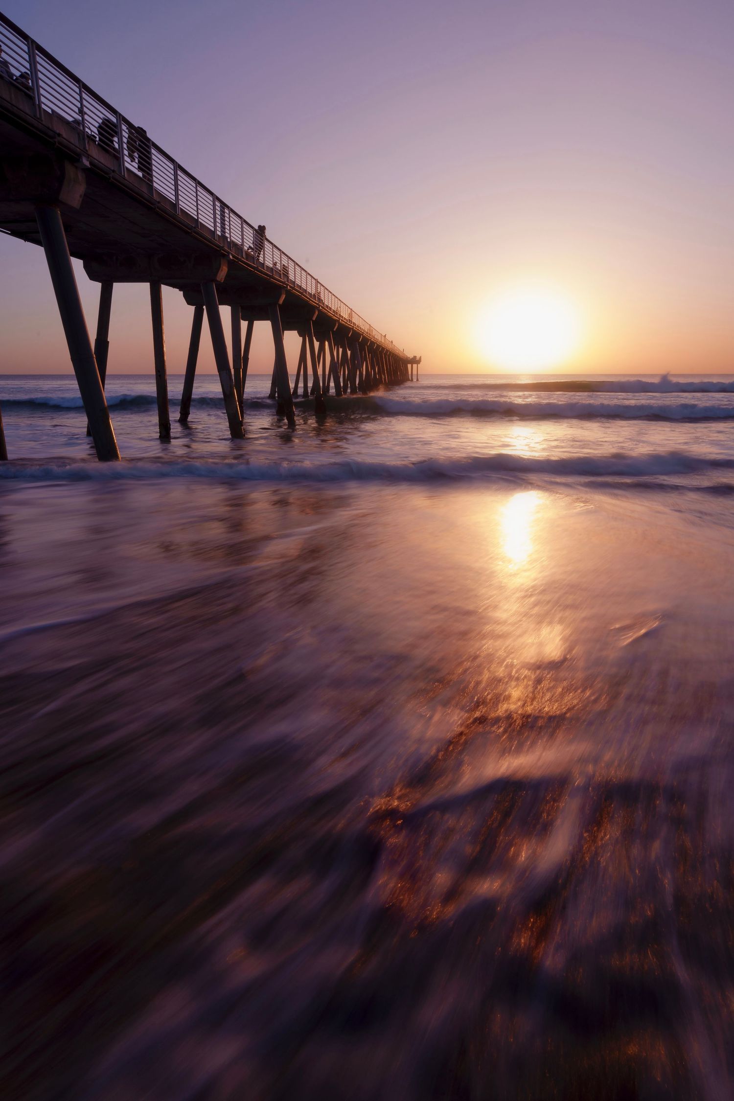 Golden sunset reflects on wet sand beneath a wooden pier as ocean waves rush toward shore.