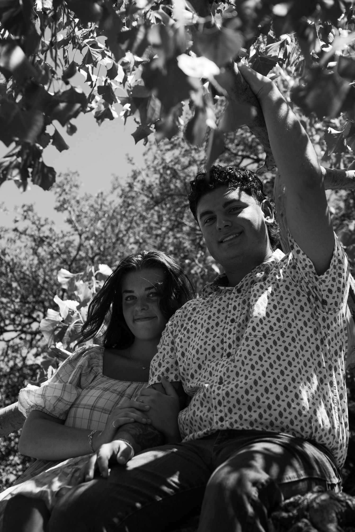 Black and white photo of couple sitting together under leafy tree branches.