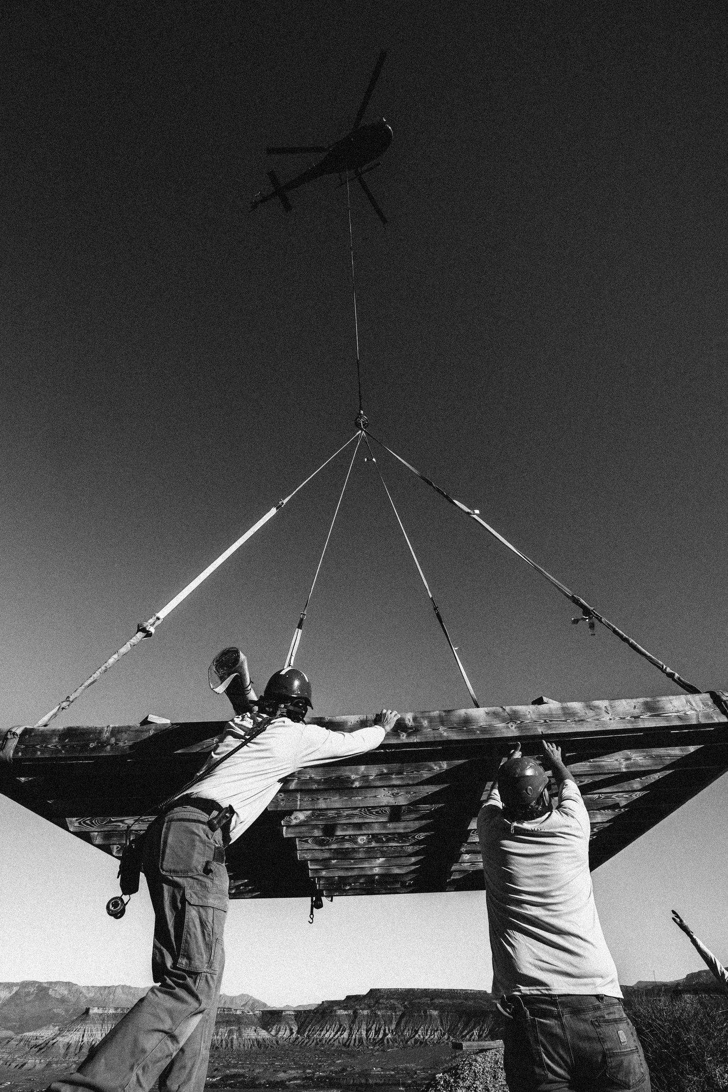 Black and white photo of workers guiding a wooden platform suspended from a helicopter in a canyon.