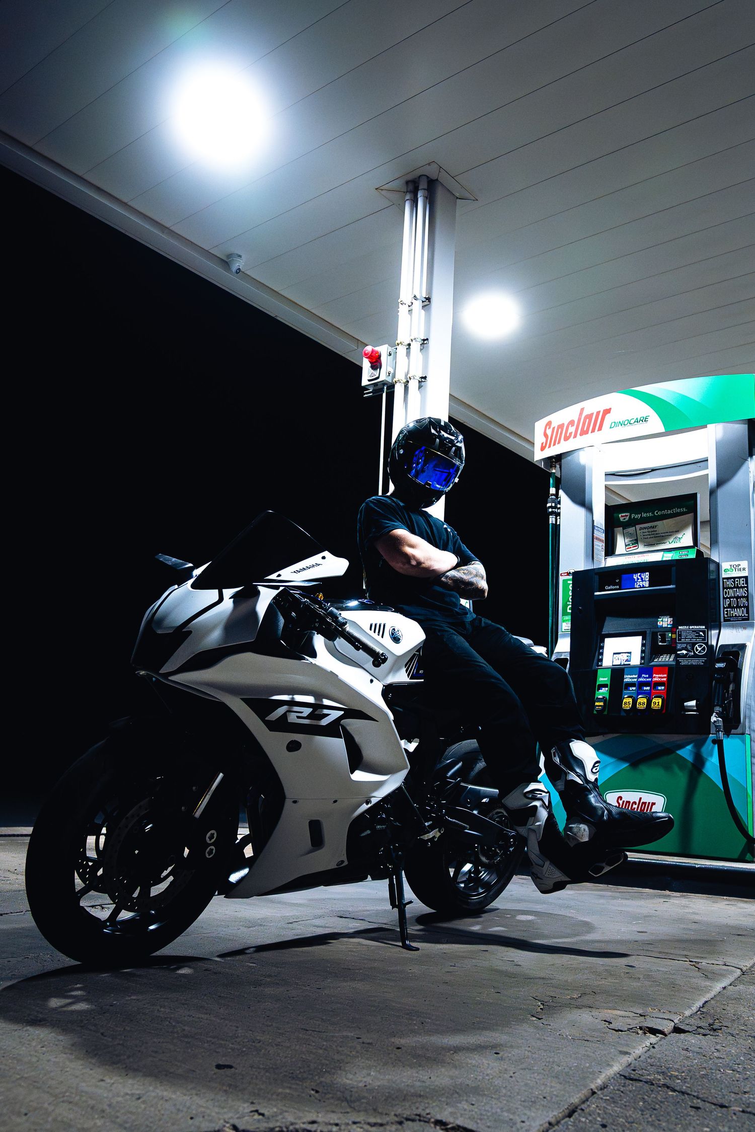 Motorcyclist in helmet poses on white Yamaha R7 sport bike at Sinclair gas station at night.