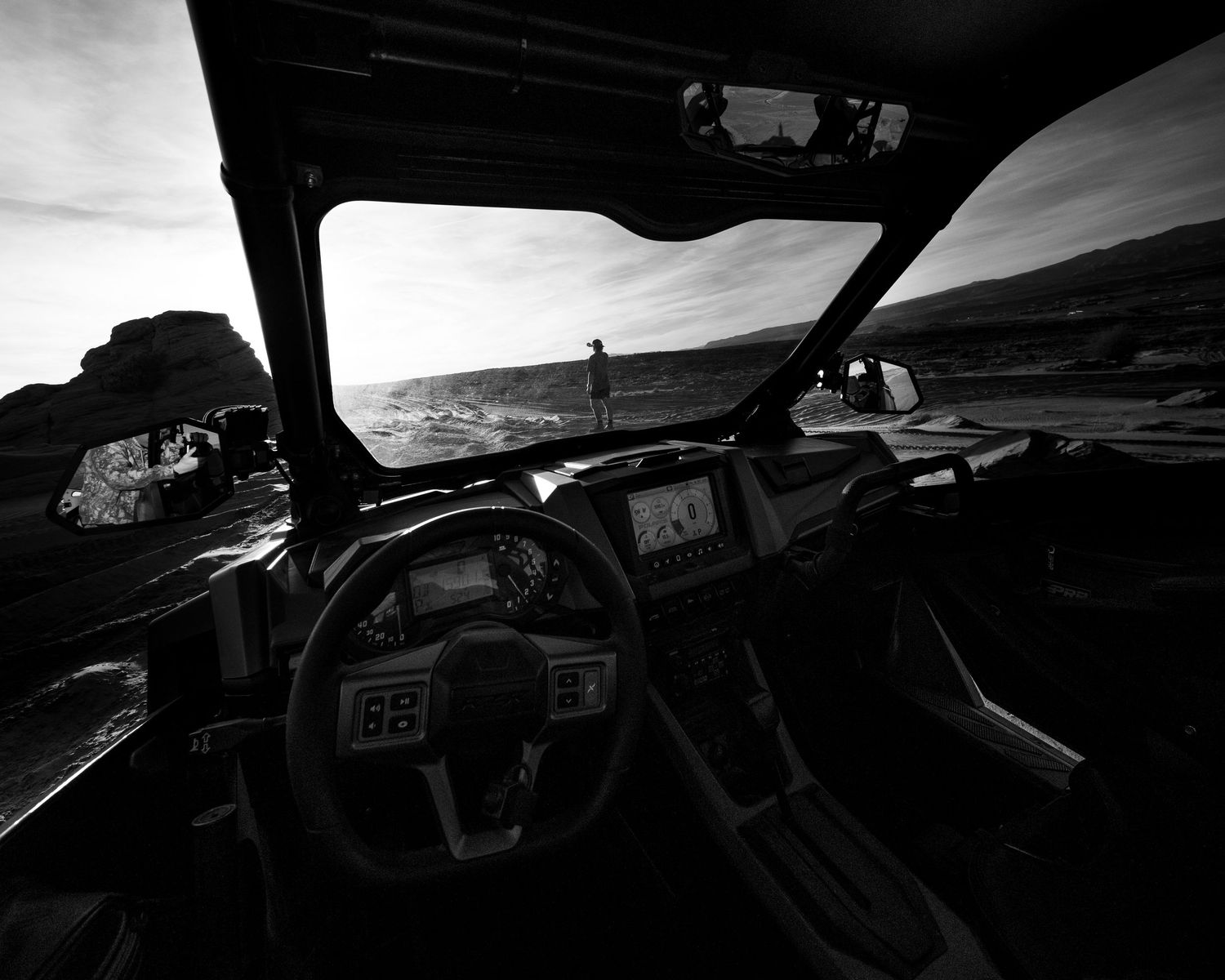 Black and white interior cockpit view of a UTV overlooking a rocky coastal shoreline at sunset.