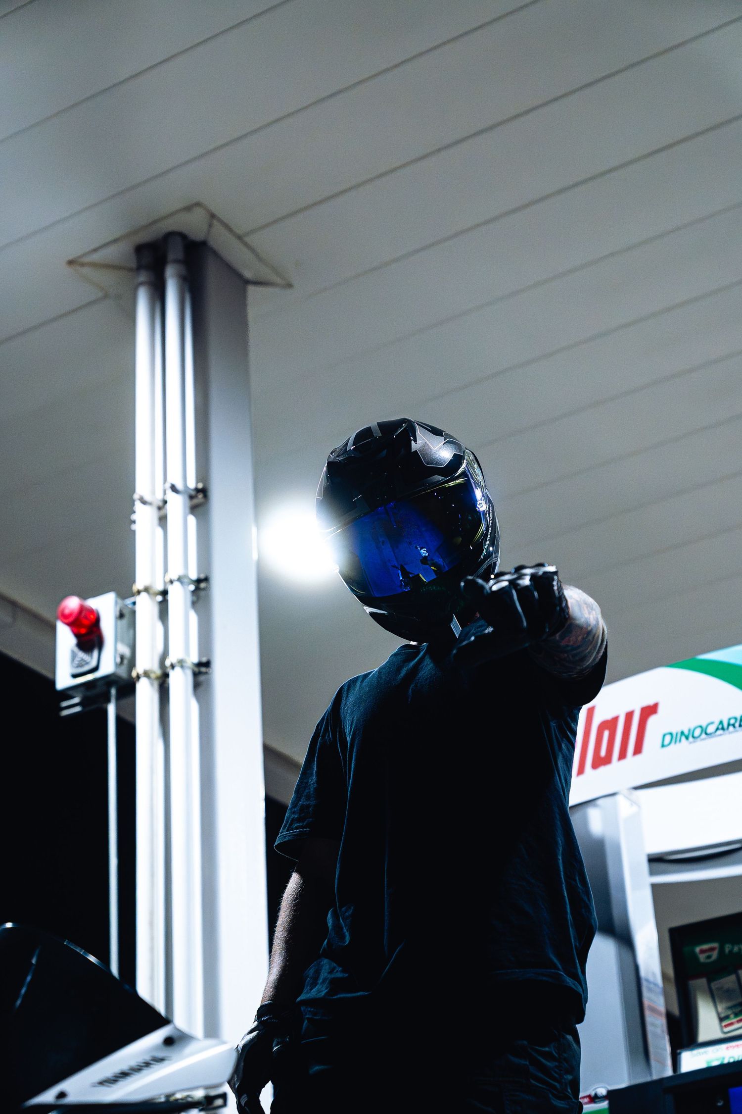 Motorcyclist in black helmet with blue visor stands at Sinclair Dinocare gas station at night.