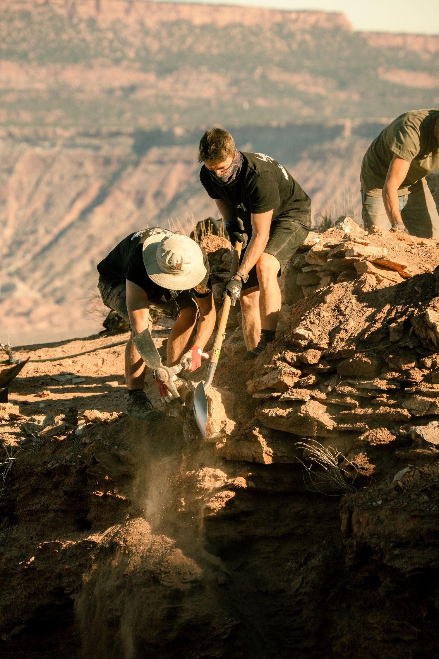 Two workers in black shirts digging with shovels on a rocky canyon cliff edge in desert terrain.