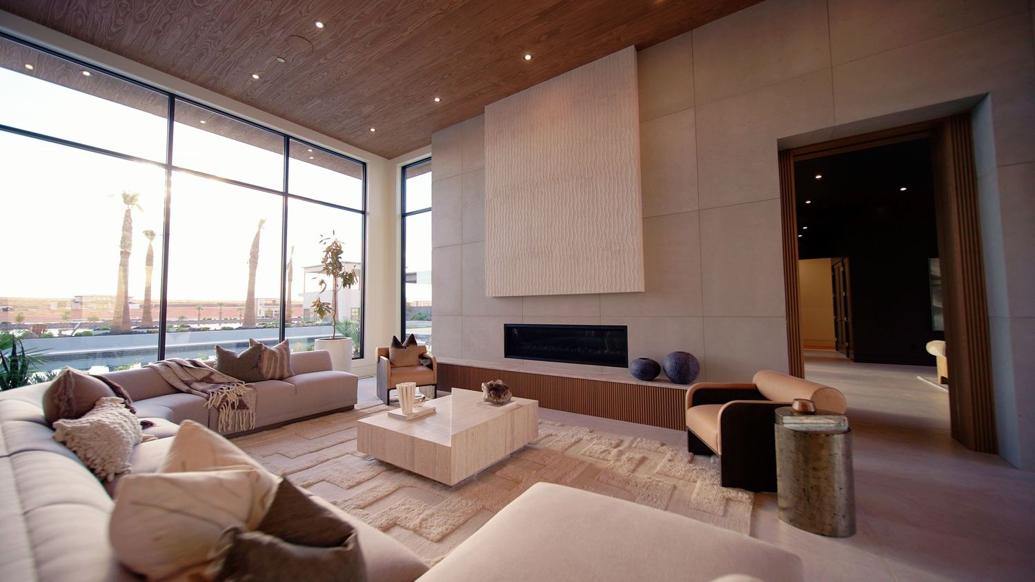 Luxury living room with floor-to-ceiling windows, modern fireplace, neutral tones, and elegant furnishings overlooking a scenic view.