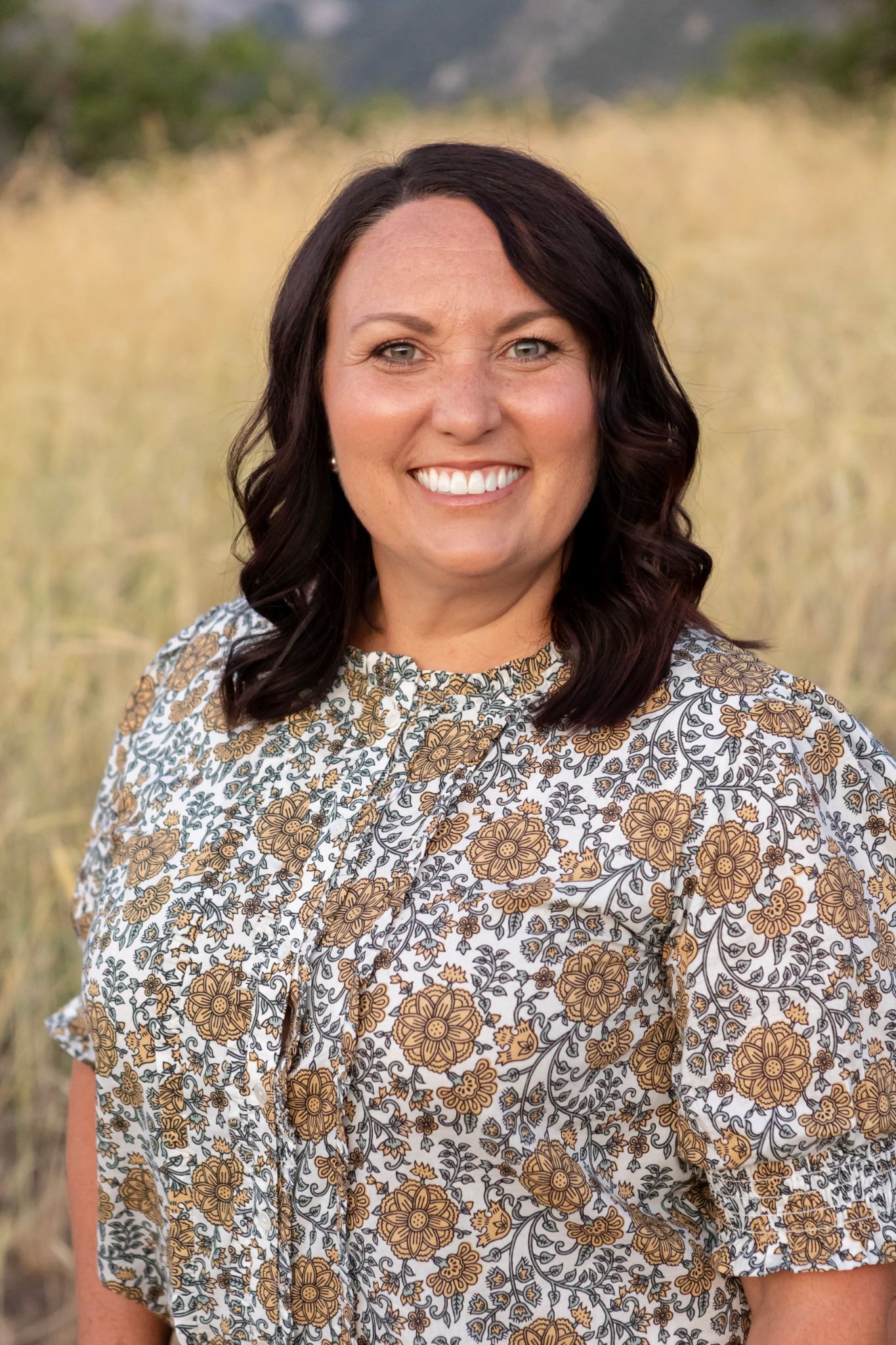 Smiling woman with dark hair wearing a floral blouse, posing outdoors in a golden field.