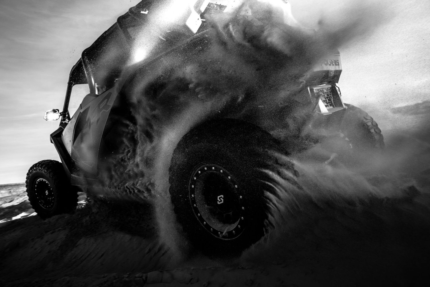Black and white dramatic low-angle shot of a UTV kicking up sand on desert dunes.