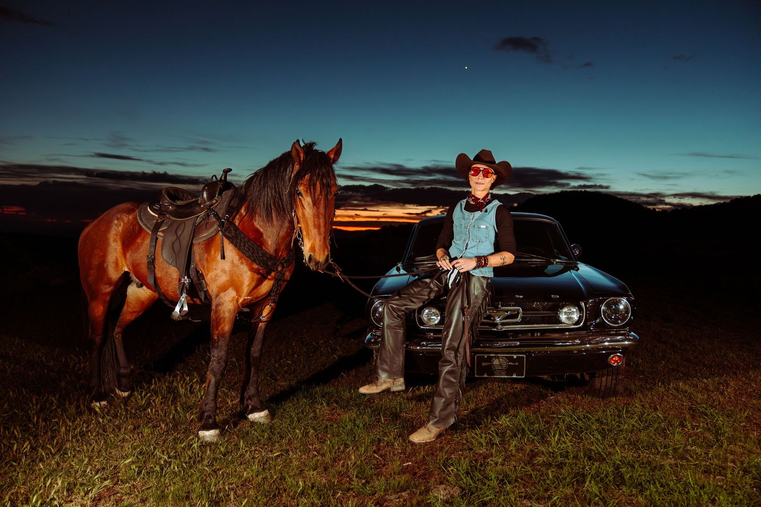 Cowboy in hat leans against a classic car beside his brown horse at colorful sunset.