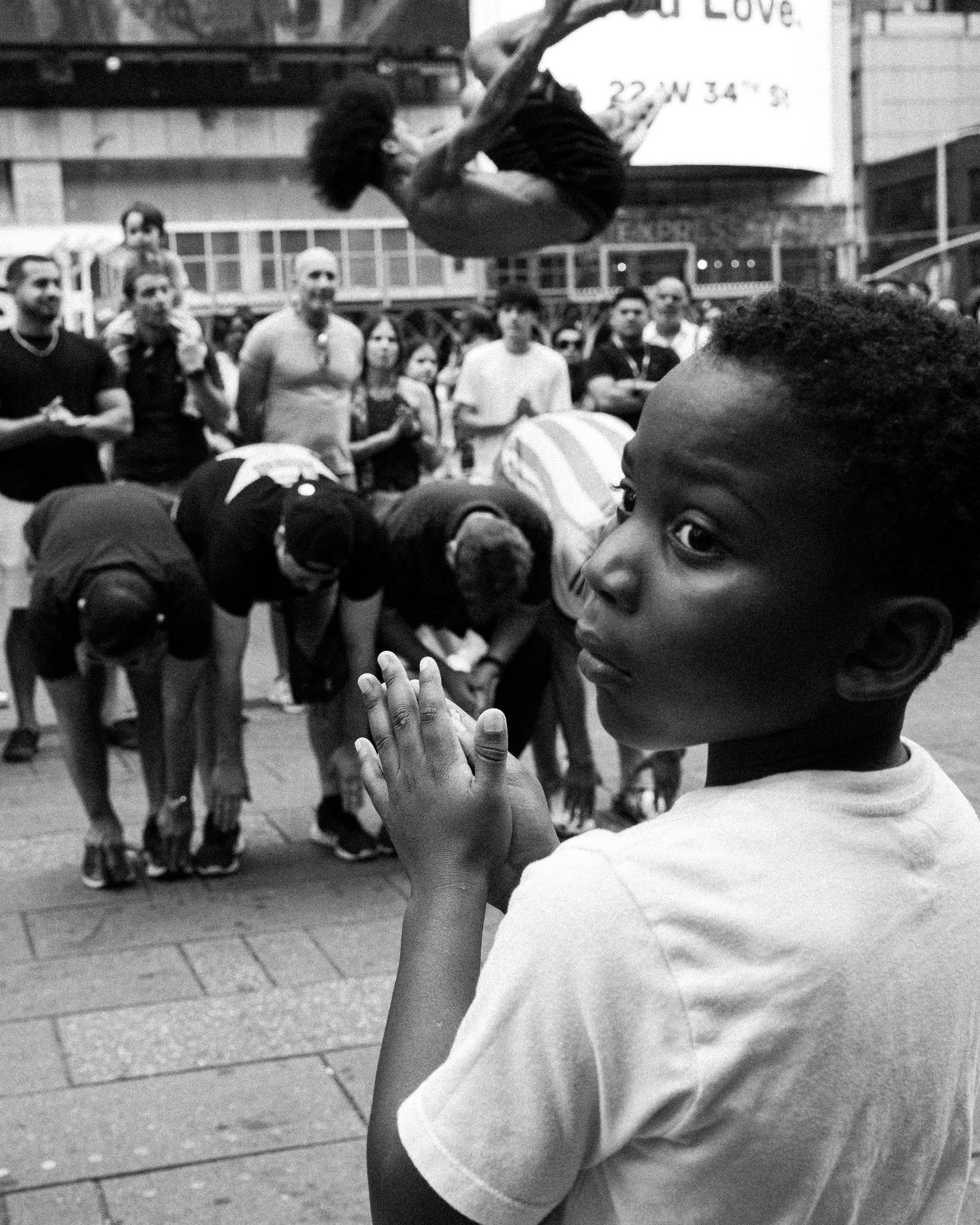 Black and white photo of a young boy clasping hands while watching street performers in a busy city square.