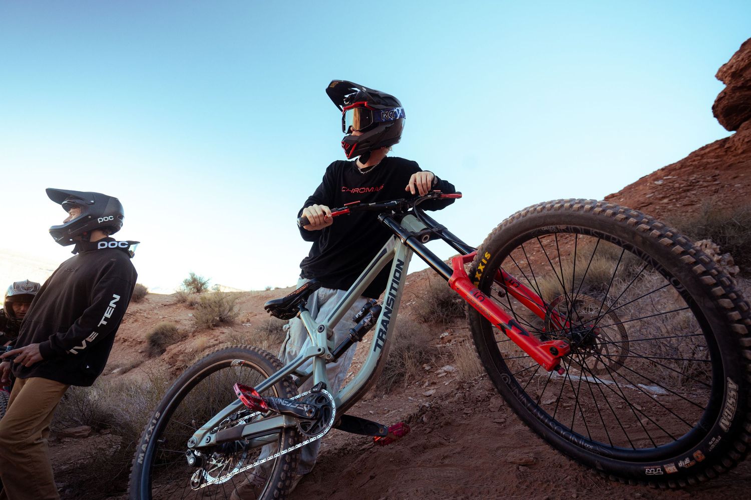 Two mountain bikers in helmets rest with Transition bike on rocky desert terrain under blue sky.
