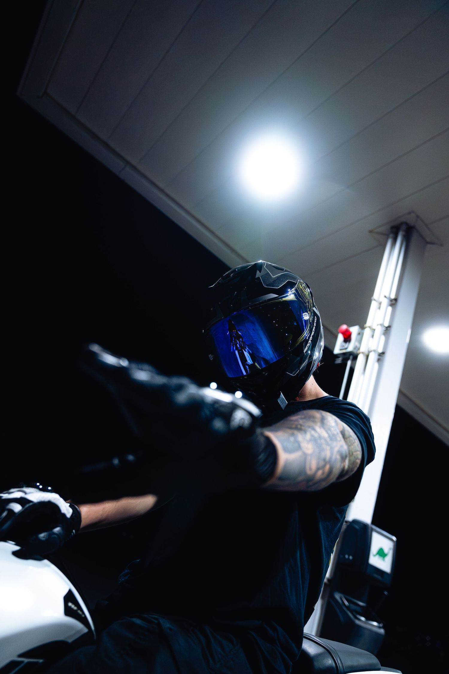 Tattooed motorcyclist in black helmet with blue visor rides white sport bike in gas station.