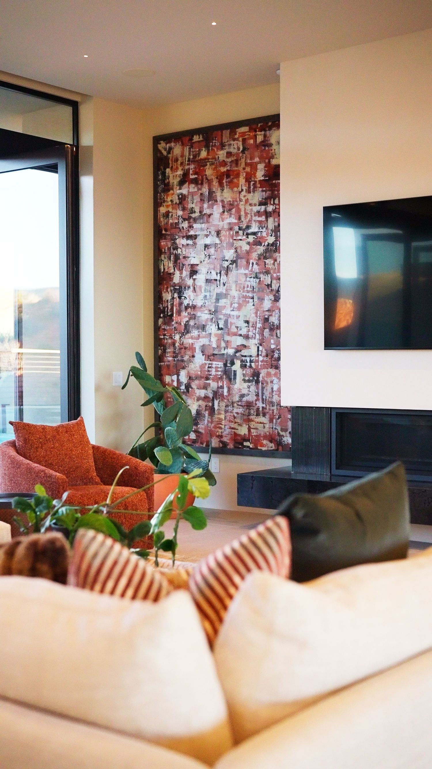 Modern living room with abstract red artwork, wall-mounted TV, fireplace, orange armchair, and lush green plants.