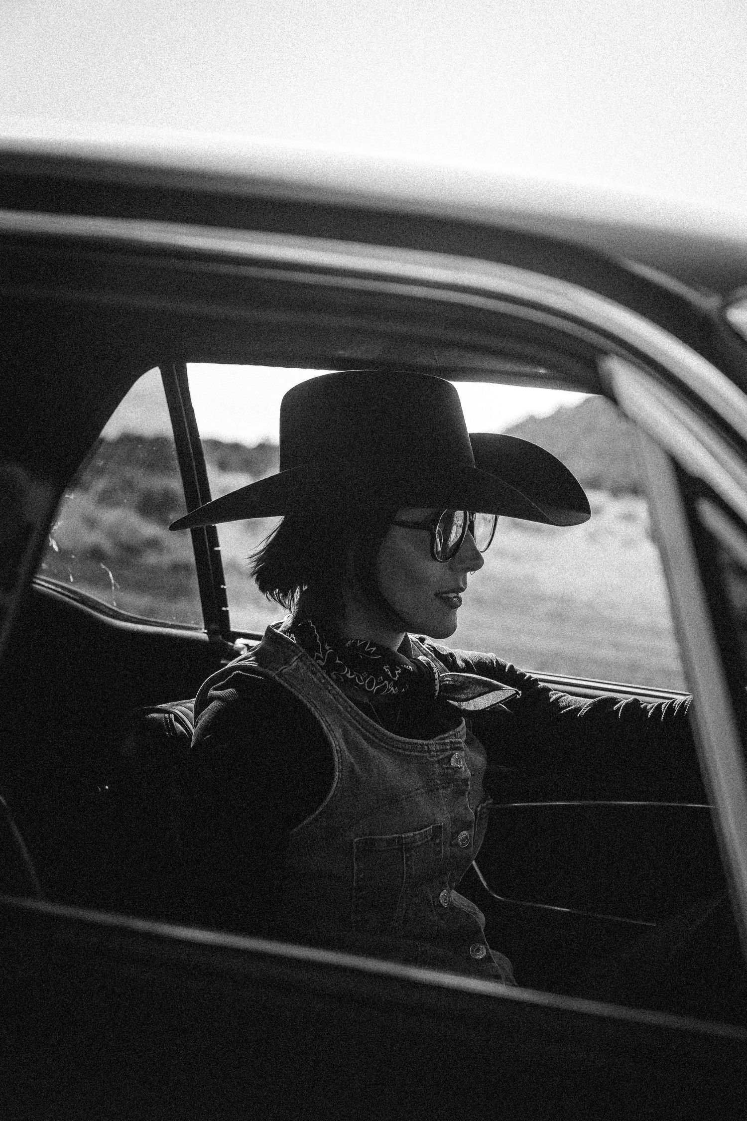 Black and white portrait of a woman in a cowboy hat and glasses sitting inside a vintage car on a desert road.